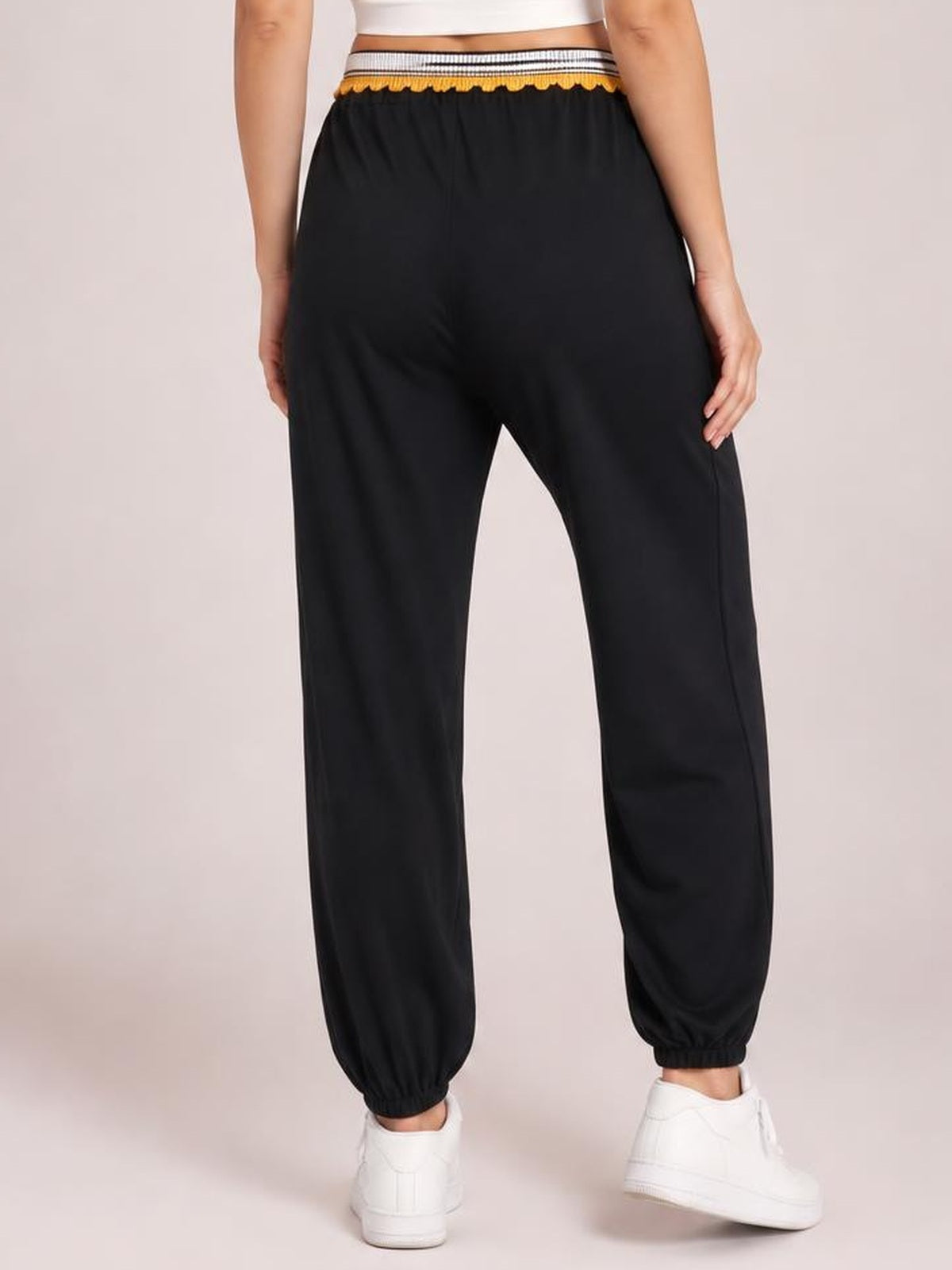 A model stands in a rear view wearing black contrast joggers with a striped elastic waistband, relaxed straight-leg silhouette, and gathered ankle cuffs.