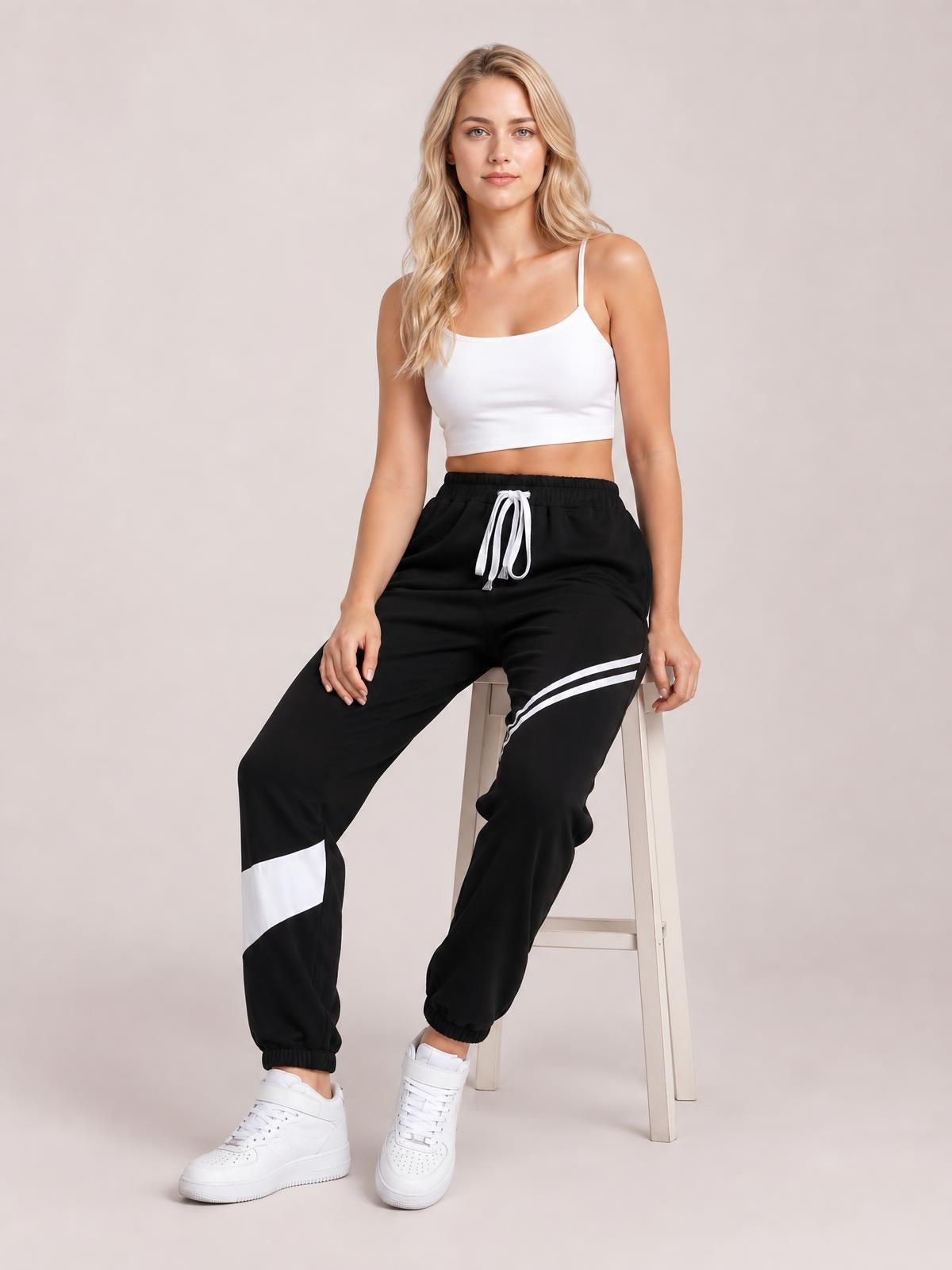 A model sits wearing black drawstring joggers with white contrast stripes, a relaxed tapered silhouette, elastic cuffs, and side pockets.