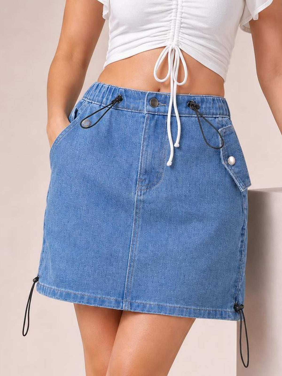Model wearing a light blue denim skirt with a straight mini silhouette, drawstring waist, buttoned front, spacious pockets, and adjustable hem ties.