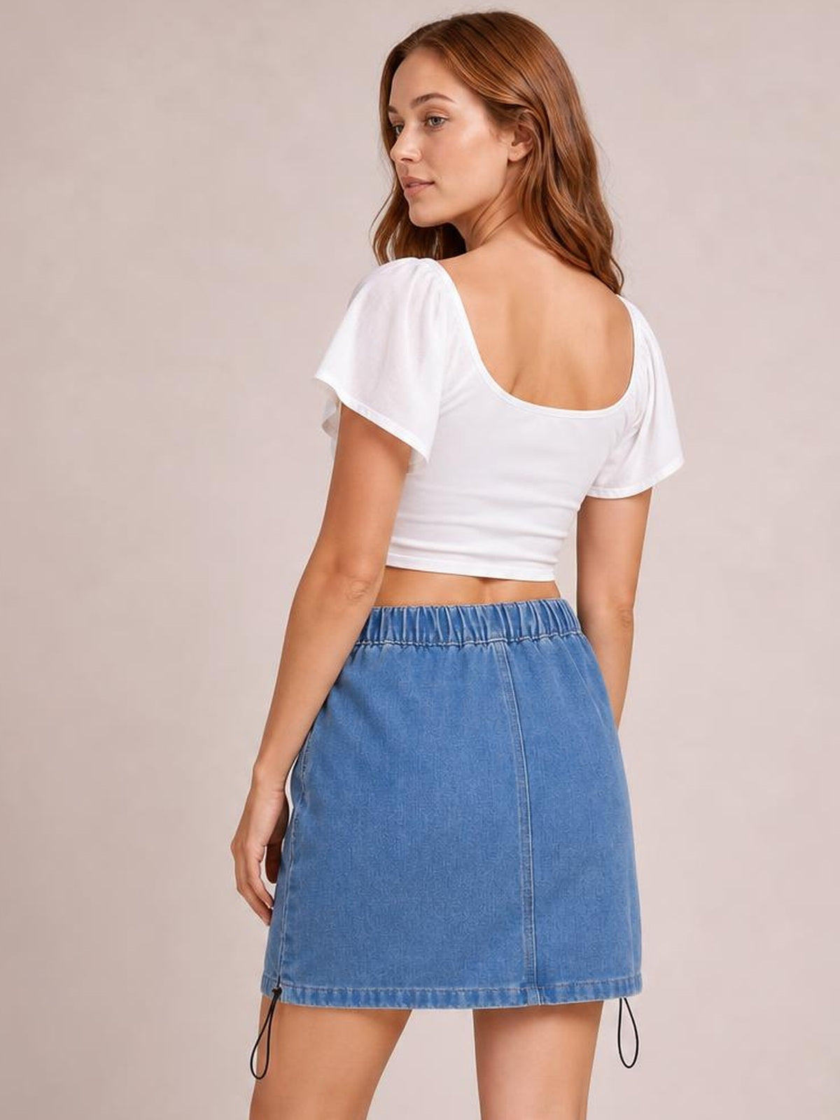 A model poses from behind wearing a light blue denim skirt with a straight mini silhouette, gathered drawstring waistband, center seam, and adjustable hem ties.