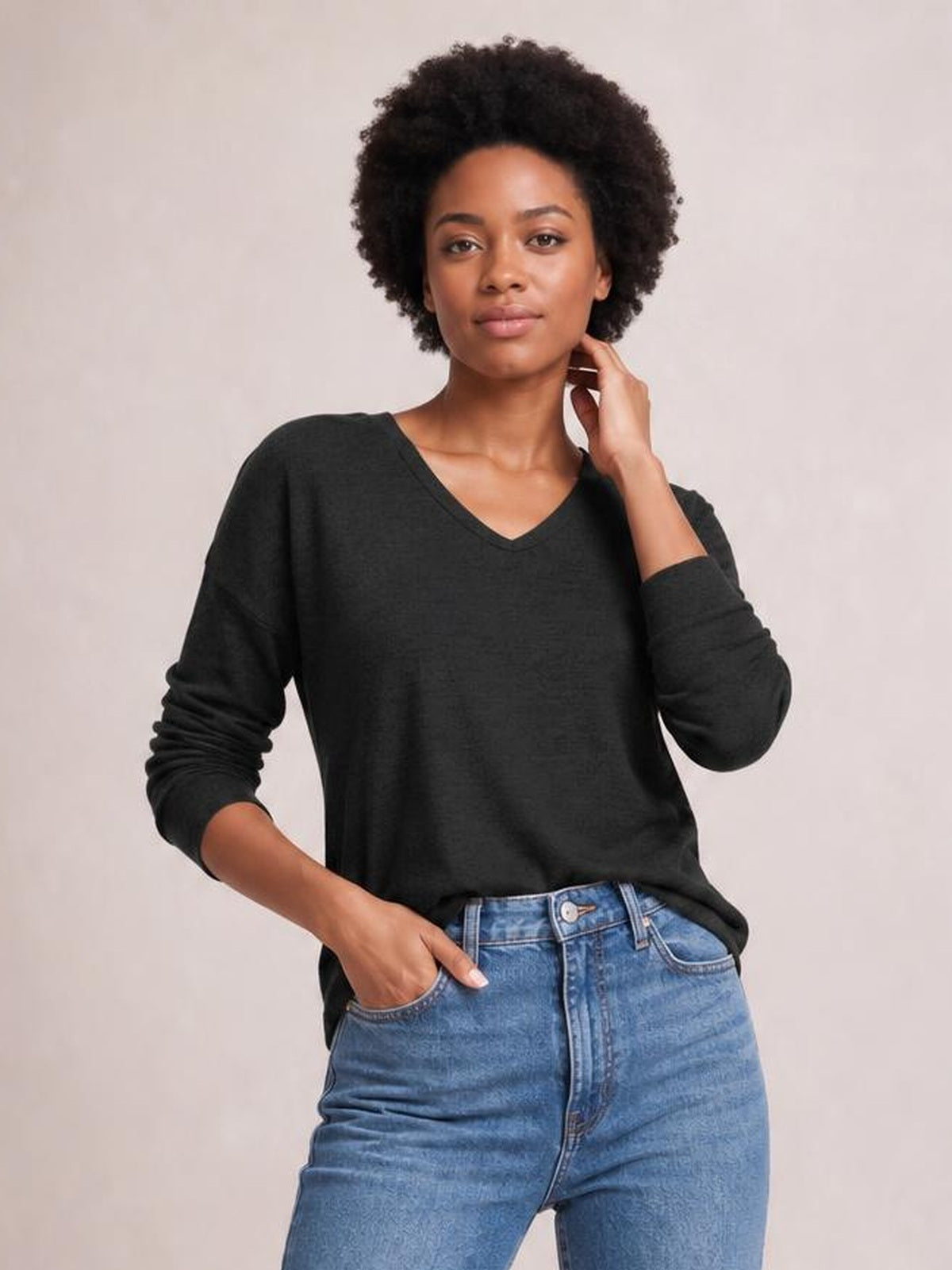 A model wears a relaxed black long-sleeve T-shirt with dropped shoulders and a V-neck, posing with one hand in her jeans pocket and the other at her neck.