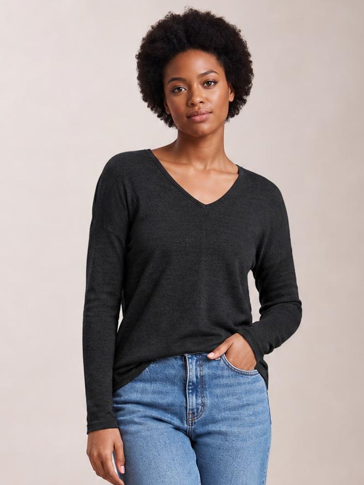 A model wears a charcoal long-sleeve T-shirt with a relaxed dropped-shoulder silhouette and V-neckline, posing with one hand in her jeans pocket.