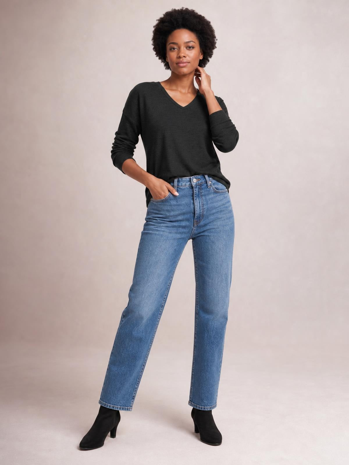 A model wears a black relaxed-fit T-shirt with dropped shoulders, long sleeves, and a V-neck, standing with one hand in her jeans pocket.