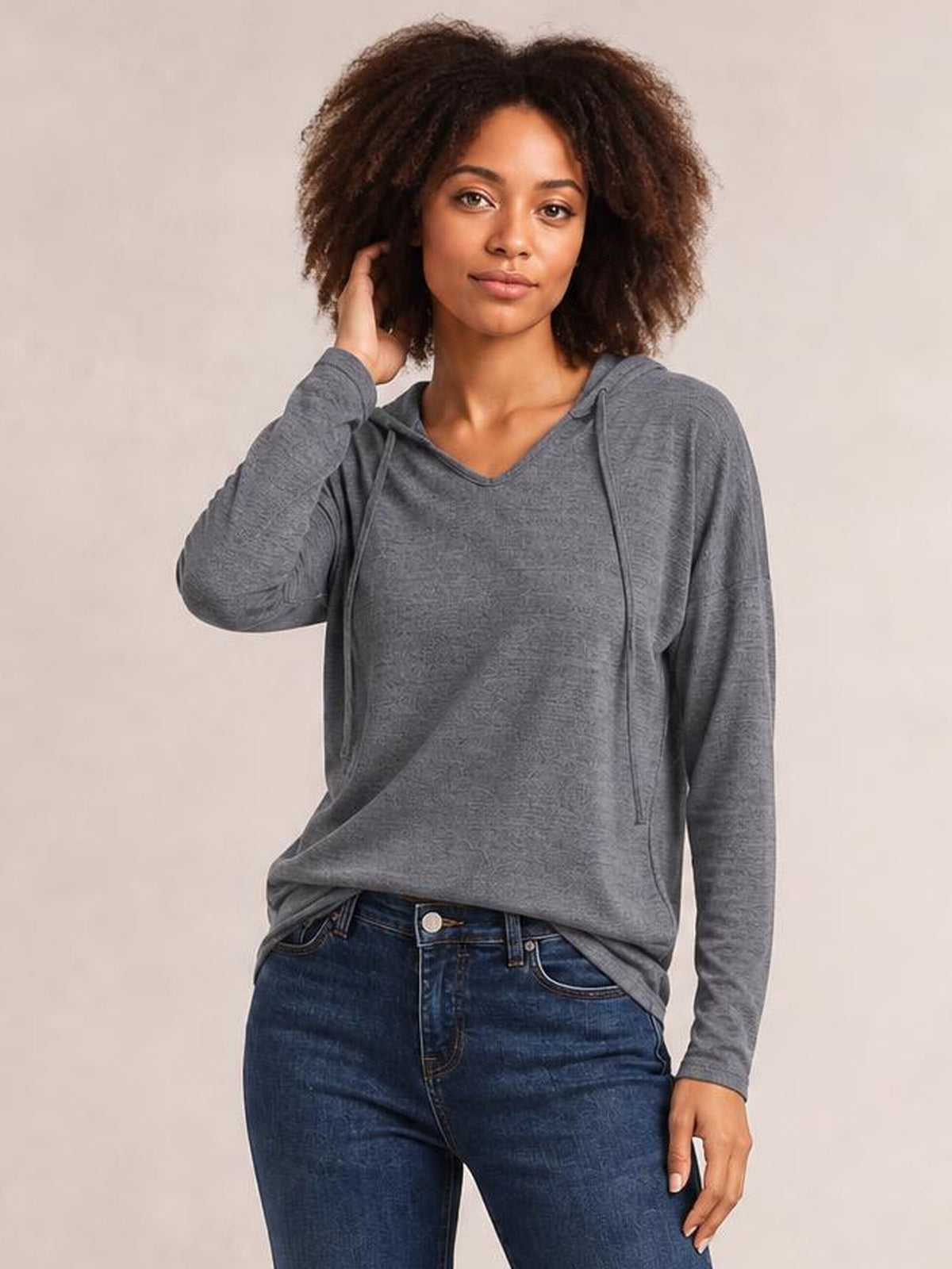 A woman poses in a relaxed gray hooded T-shirt with dropped shoulders, long sleeves, visible drawstrings, and a loose silhouette while touching her hair.