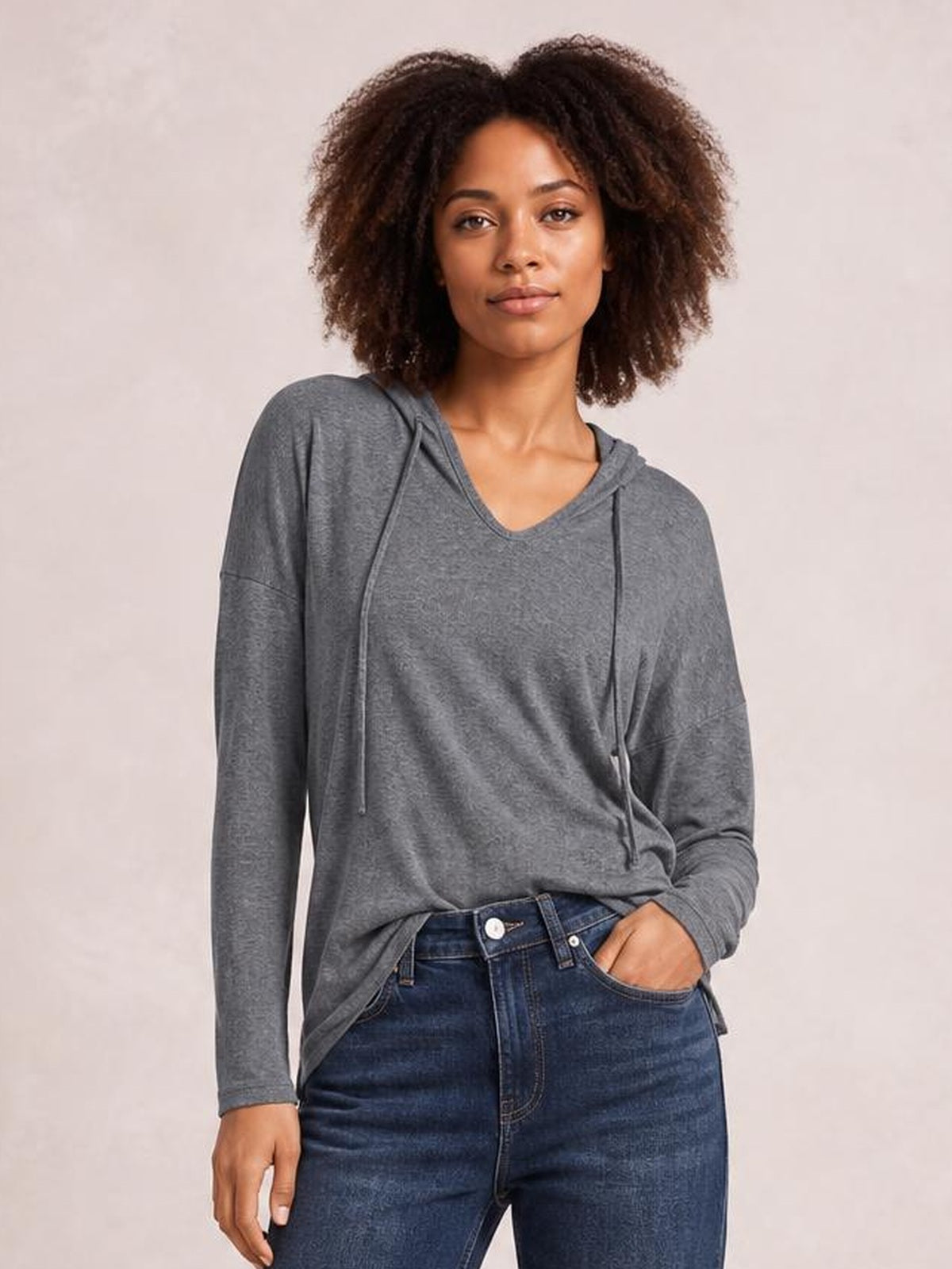 Model wears a charcoal gray drawstring dropped-shoulder hooded T-shirt with long sleeves and a relaxed silhouette, standing with one hand in her jeans pocket.