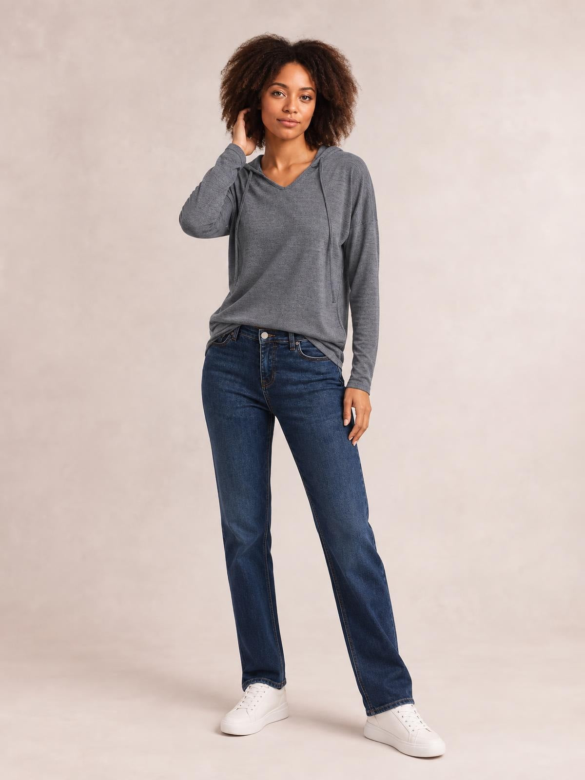 A model stands with one hand raised to her hair, wearing a relaxed gray drawstring hooded T-shirt with dropped shoulders, long sleeves, and a loose silhouette.