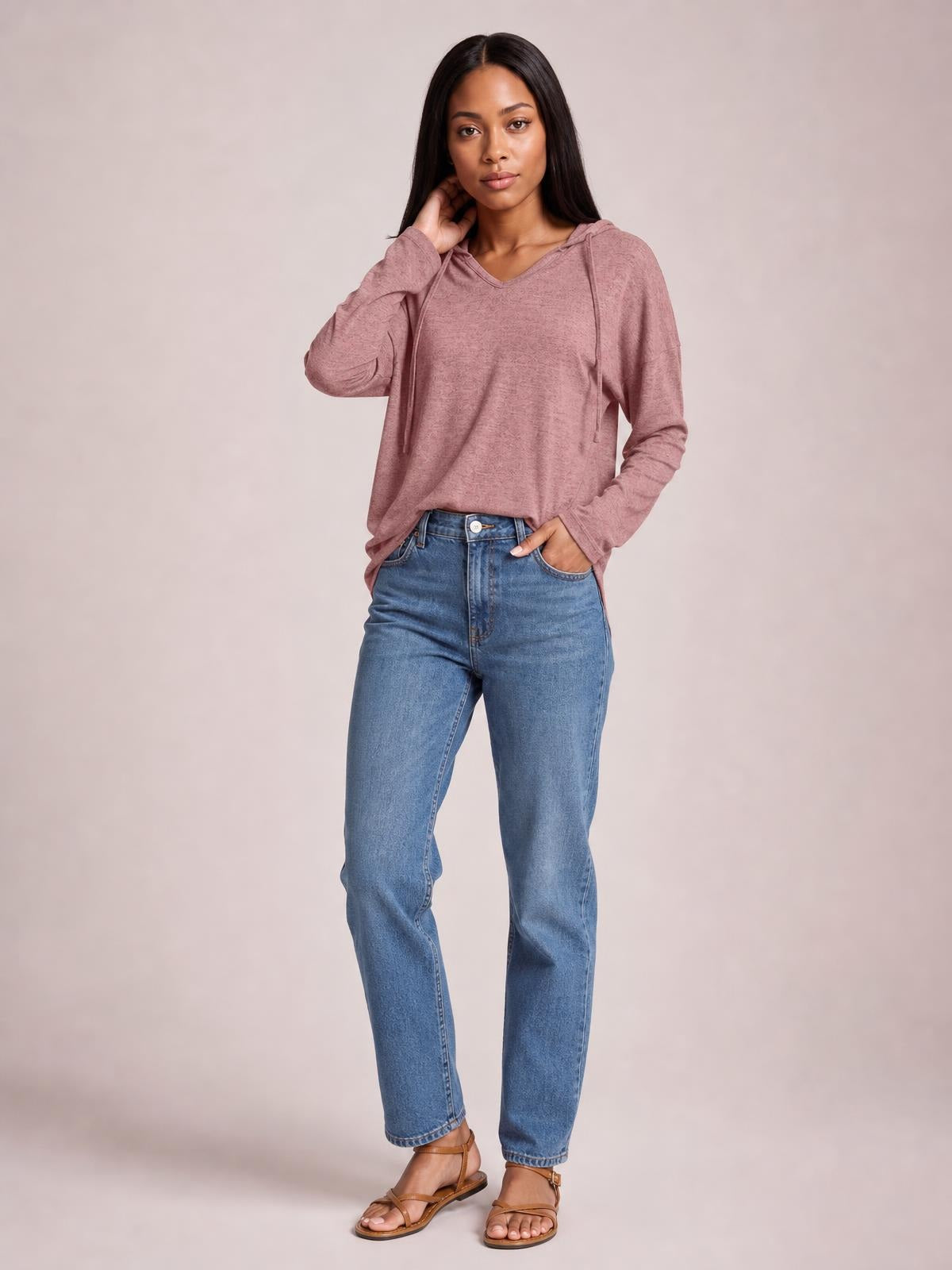 A model stands in a mauve drawstring hooded T-shirt with dropped shoulders, long sleeves, and a relaxed silhouette, touching her neck and resting one hand in her pocket.