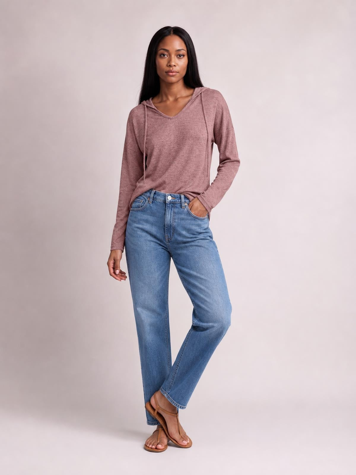 A model poses in a dusty rose drawstring hooded T-shirt with dropped shoulders, long sleeves, and a relaxed silhouette, standing with one hand in her jeans pocket.