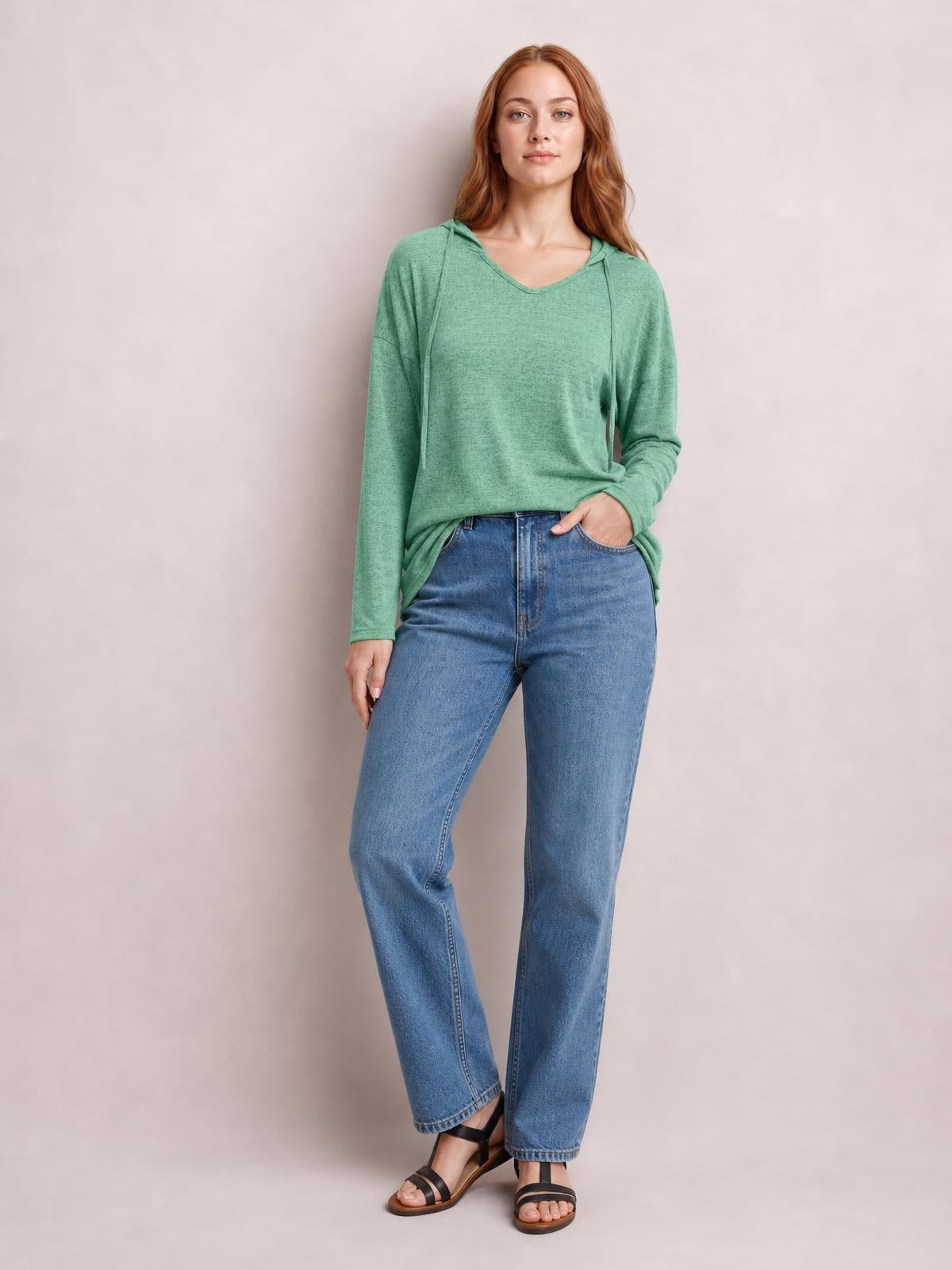 A model wears a sage green drawstring hooded T-shirt with dropped shoulders, long sleeves, and a relaxed silhouette, standing with one hand in her jeans pocket.