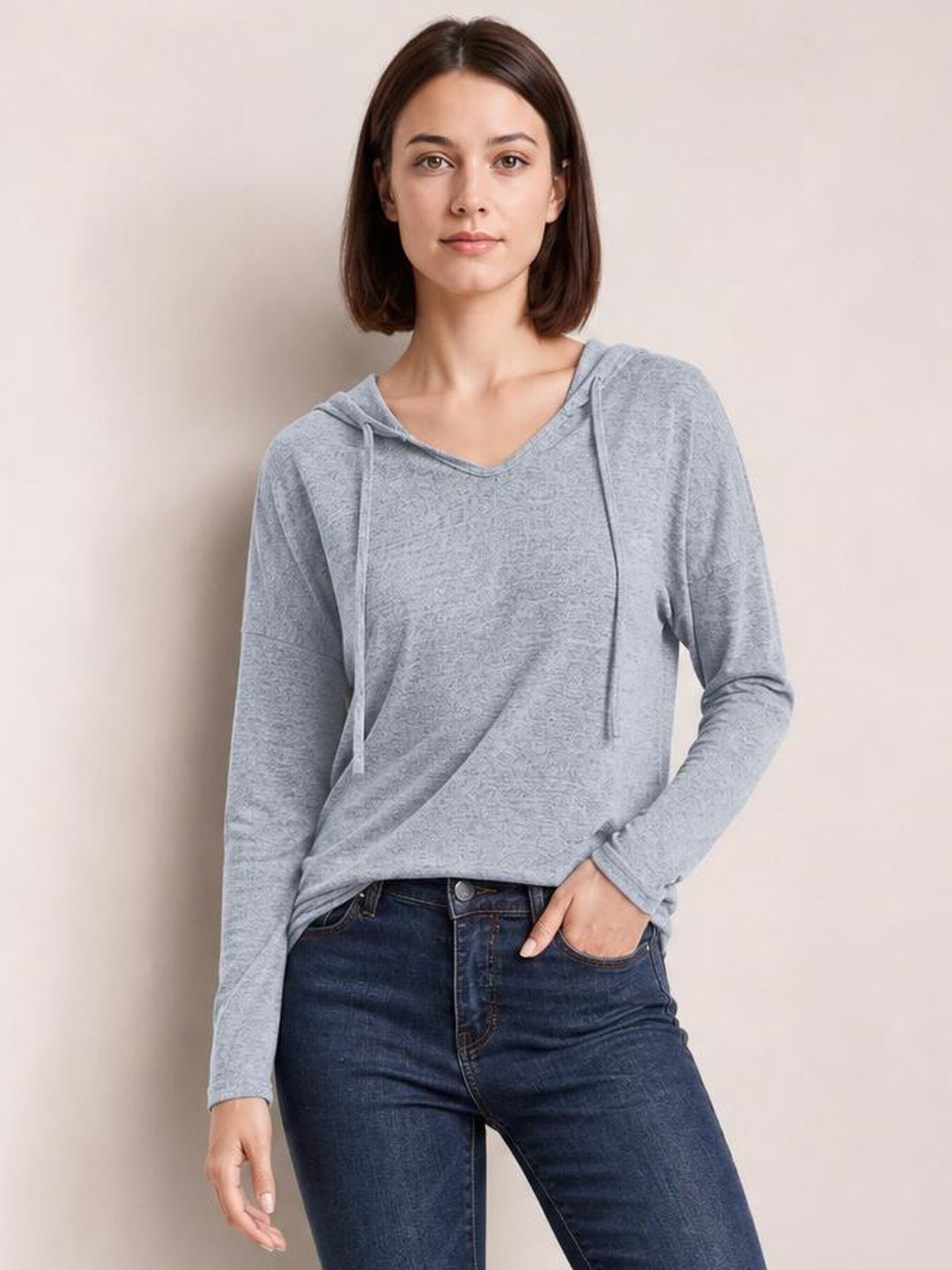 A model poses with one hand in her jeans pocket, wearing a light gray dropped-shoulder hooded T-shirt with long sleeves and front drawstrings.