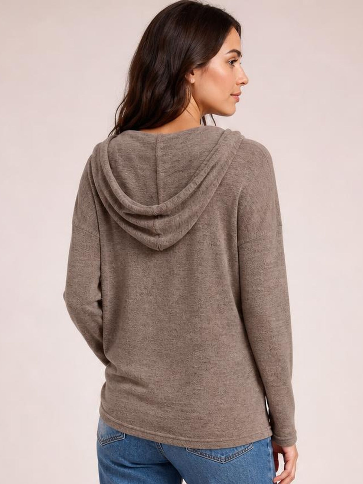 A woman turns in a three-quarter back pose wearing a taupe hooded T-shirt with a relaxed dropped-shoulder silhouette, long sleeves, and soft drape.