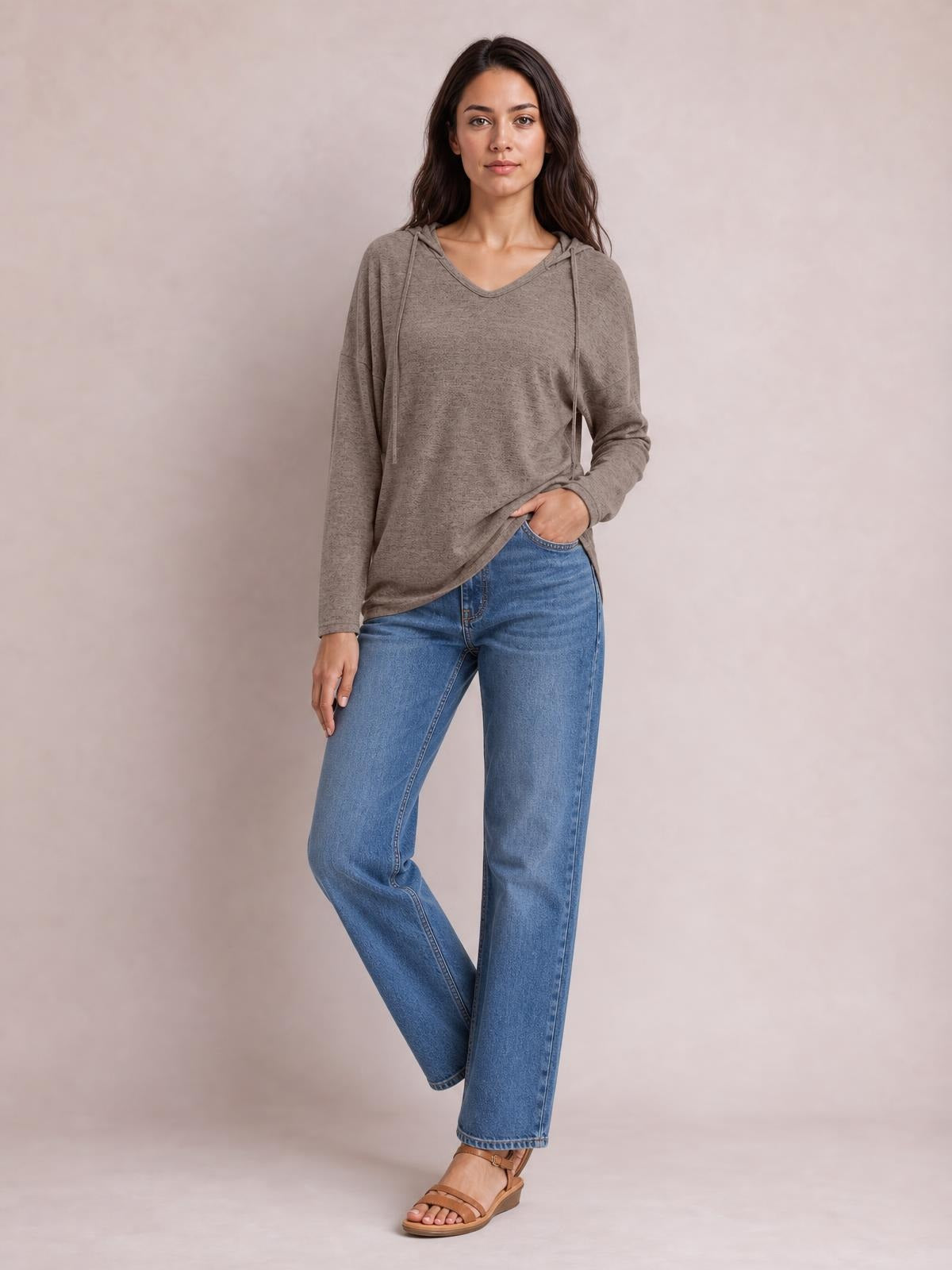 Model wears a taupe drawstring hooded T-shirt with dropped shoulders, relaxed long sleeves, a loose silhouette, and crossed legs with one hand in her pocket.