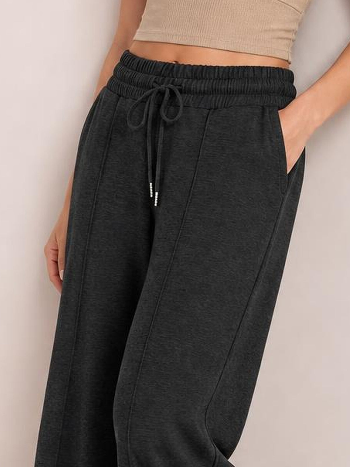 A model wears dark charcoal drawstring elastic waist wide-leg pants with roomy side pockets, visible seam detailing, and one hand tucked into a pocket.