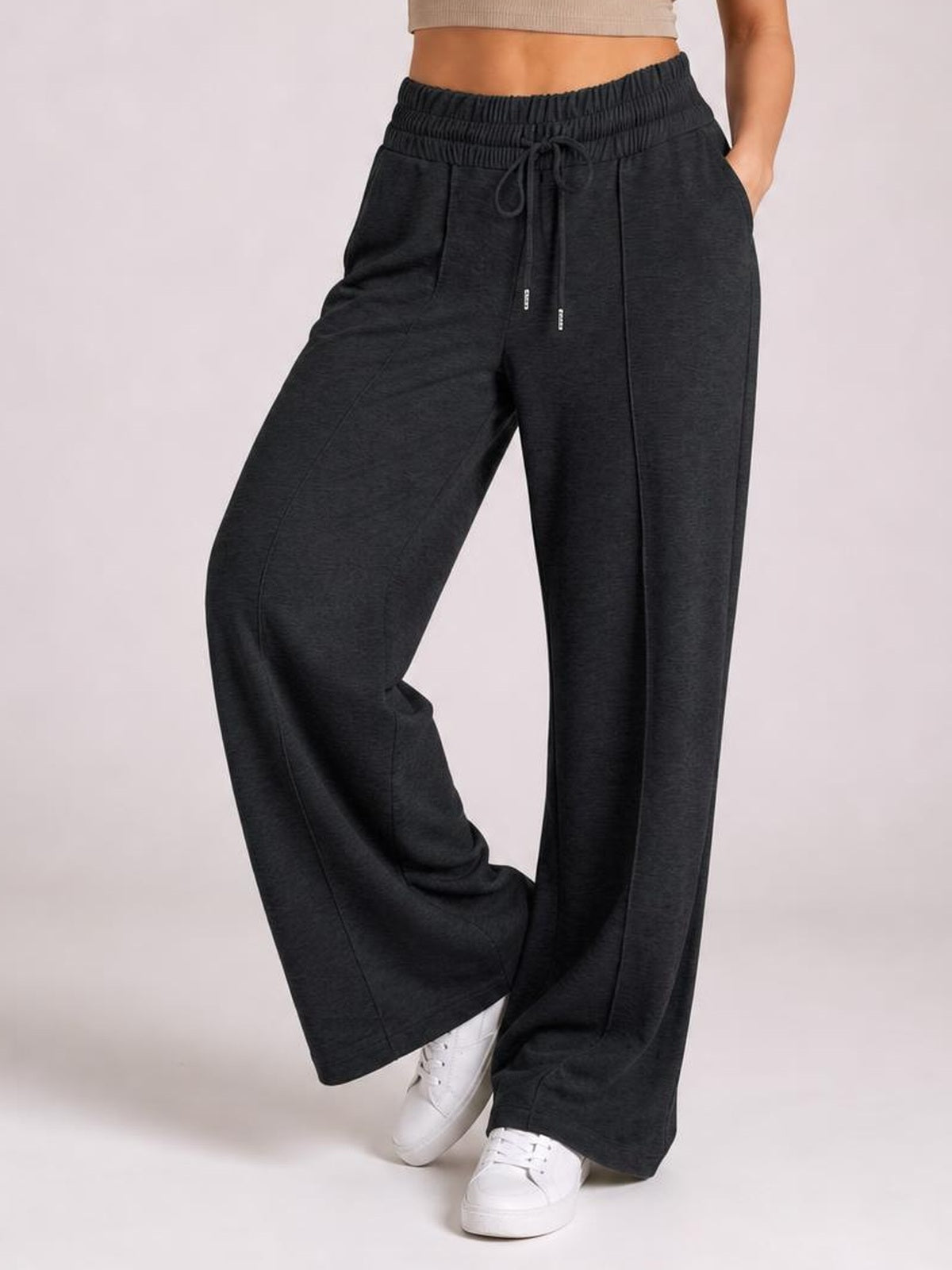 A model poses in dark wide-leg pants with a ruched elastic waist, front drawstring, pocketed construction, and relaxed flowing silhouette.