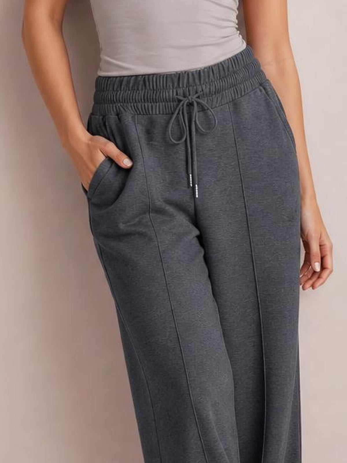 Model wearing dark gray drawstring elastic waist wide-leg pants with front pockets and stitched panel details, one hand resting in a pocket.