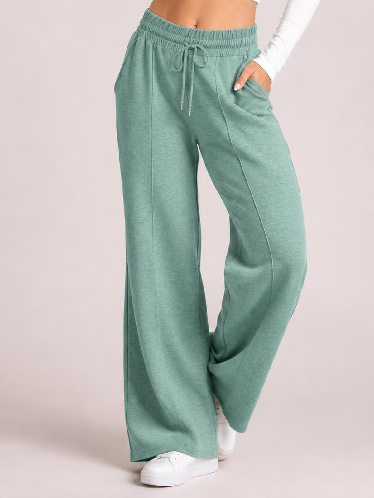 A model wears sage green drawstring elastic waist wide leg pants with front seam detailing and side pockets, standing with one hand in a pocket.