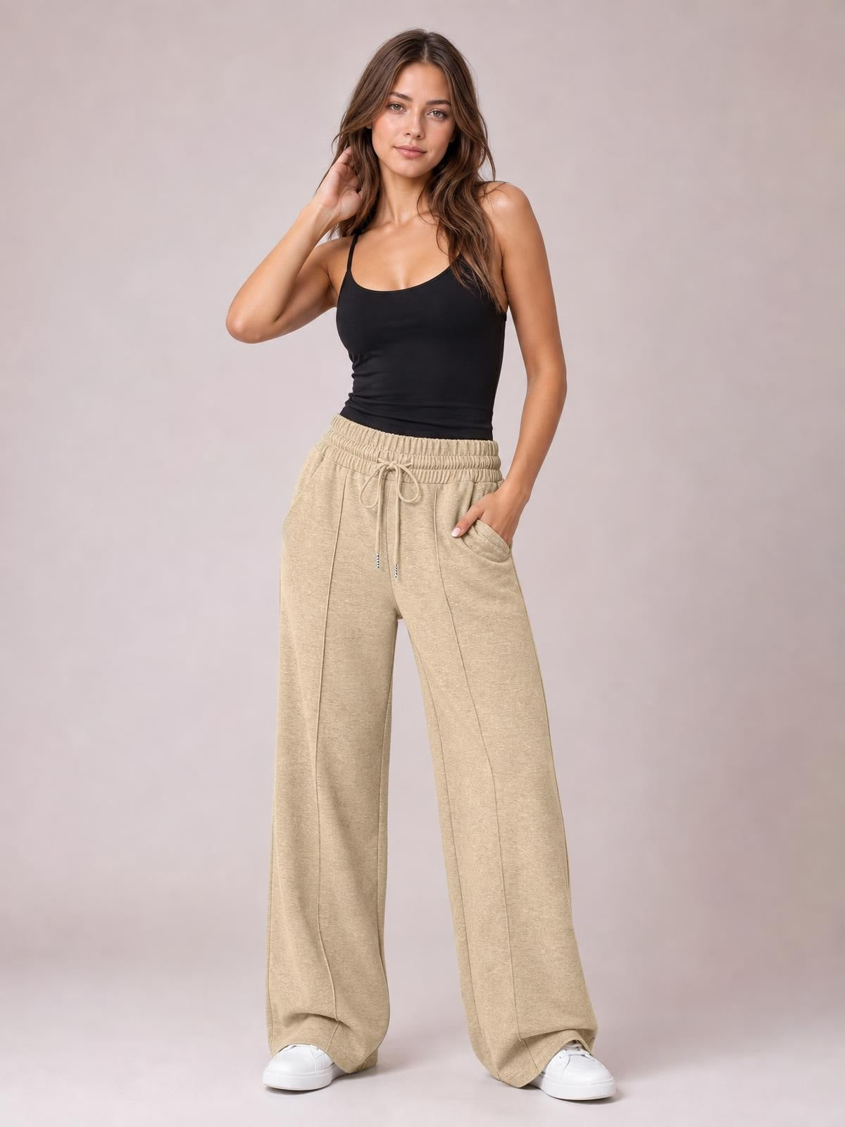 Model poses in beige drawstring elastic waist wide leg pants with front pockets, a relaxed flowy silhouette, and full-length hems over white sneakers.