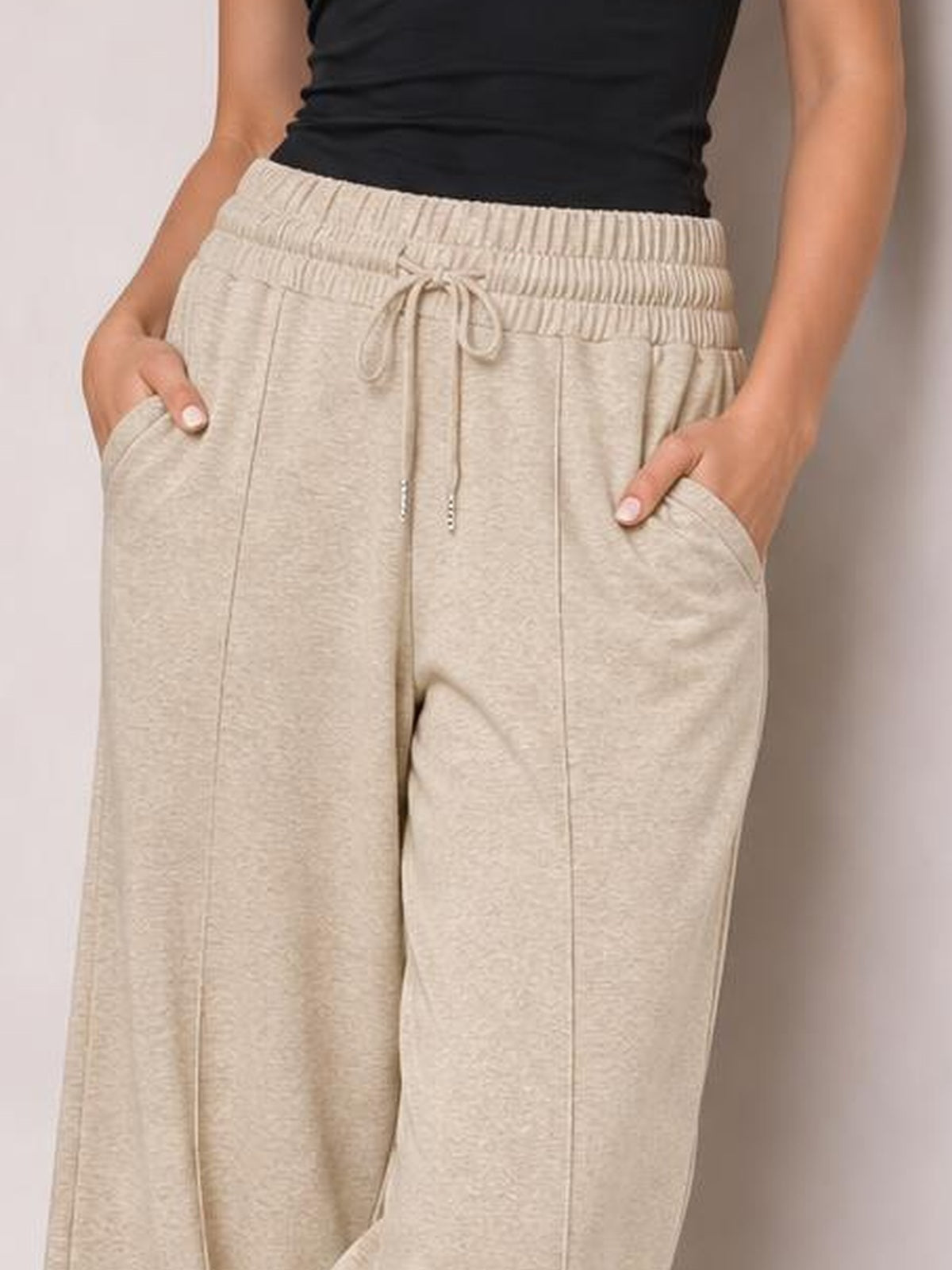 Model wearing beige drawstring elastic waist wide leg pants with front pockets and visible vertical seam detailing, posed with both hands in the pockets.
