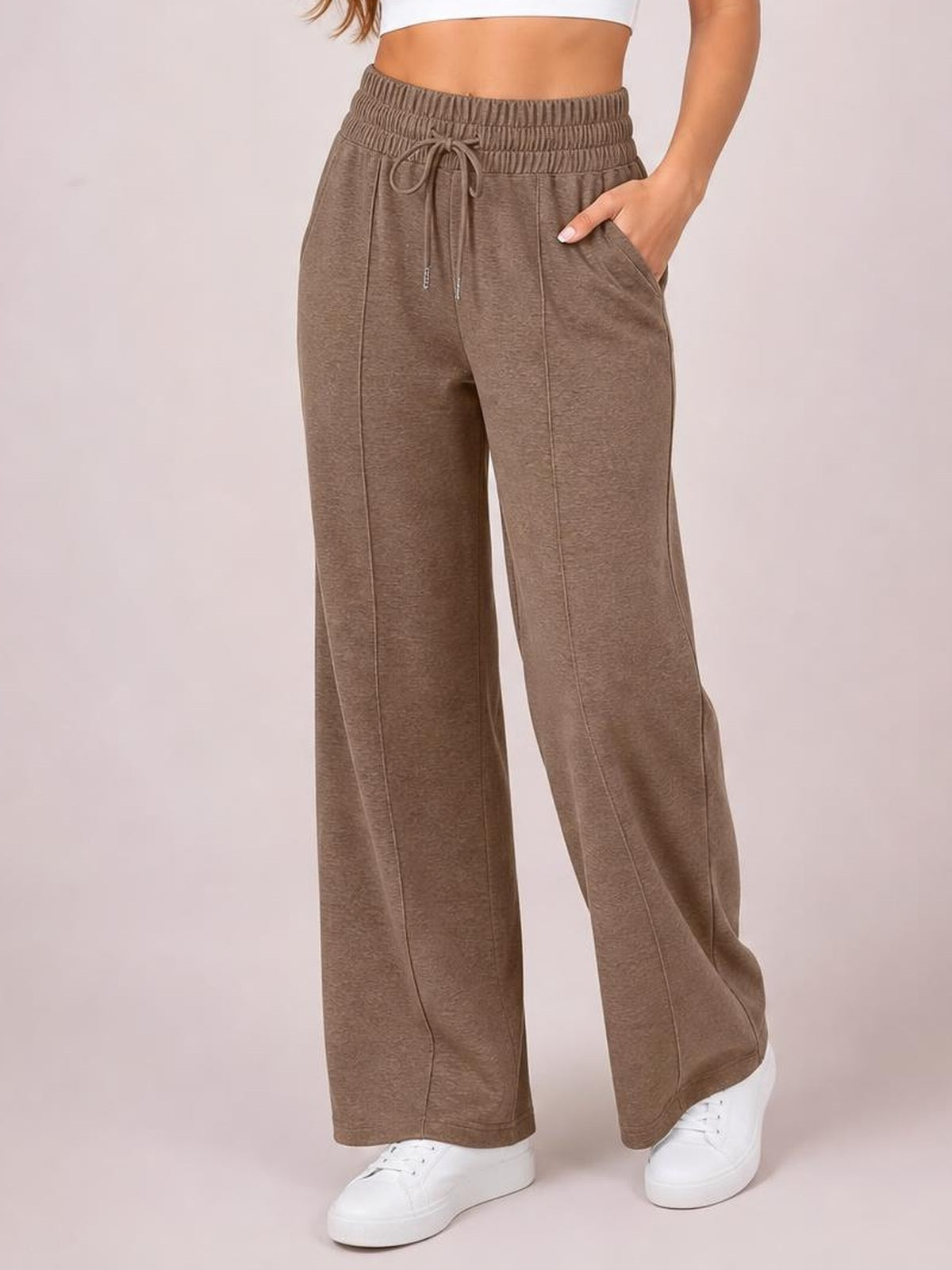 A model stands in taupe drawstring elastic-waist wide-leg pants with visible vertical seam details and side pockets, one hand tucked into a pocket.