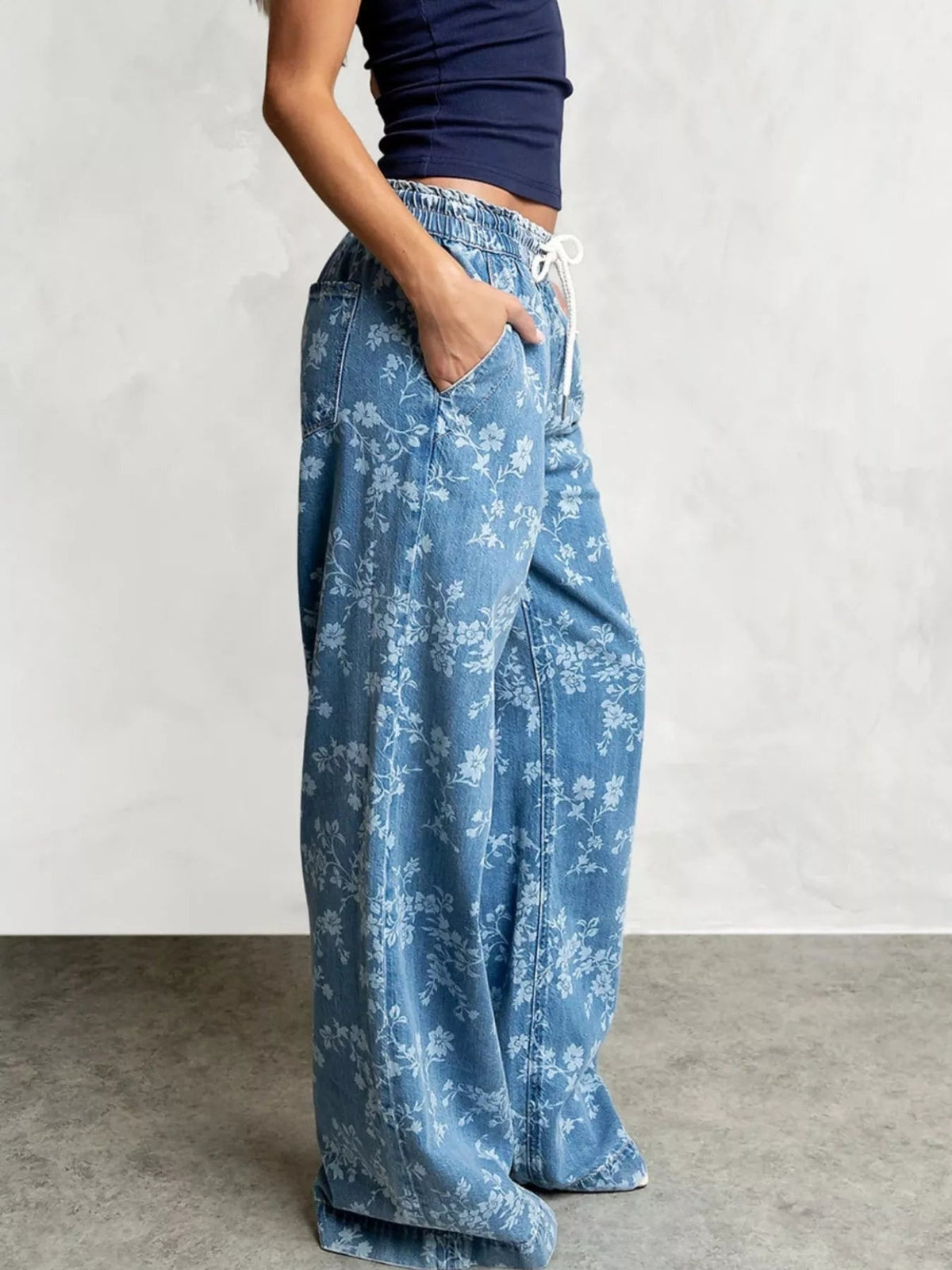 Model poses in light blue washed denim jeans with a white floral print, elastic drawstring waist, pockets, and a flowing wide-leg silhouette.