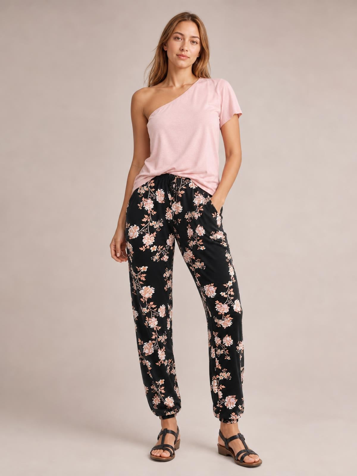 Model wearing black floral drawstring pants with side pockets, a relaxed straight-leg silhouette, and elasticized ankle cuffs, posed with one hand in a pocket.