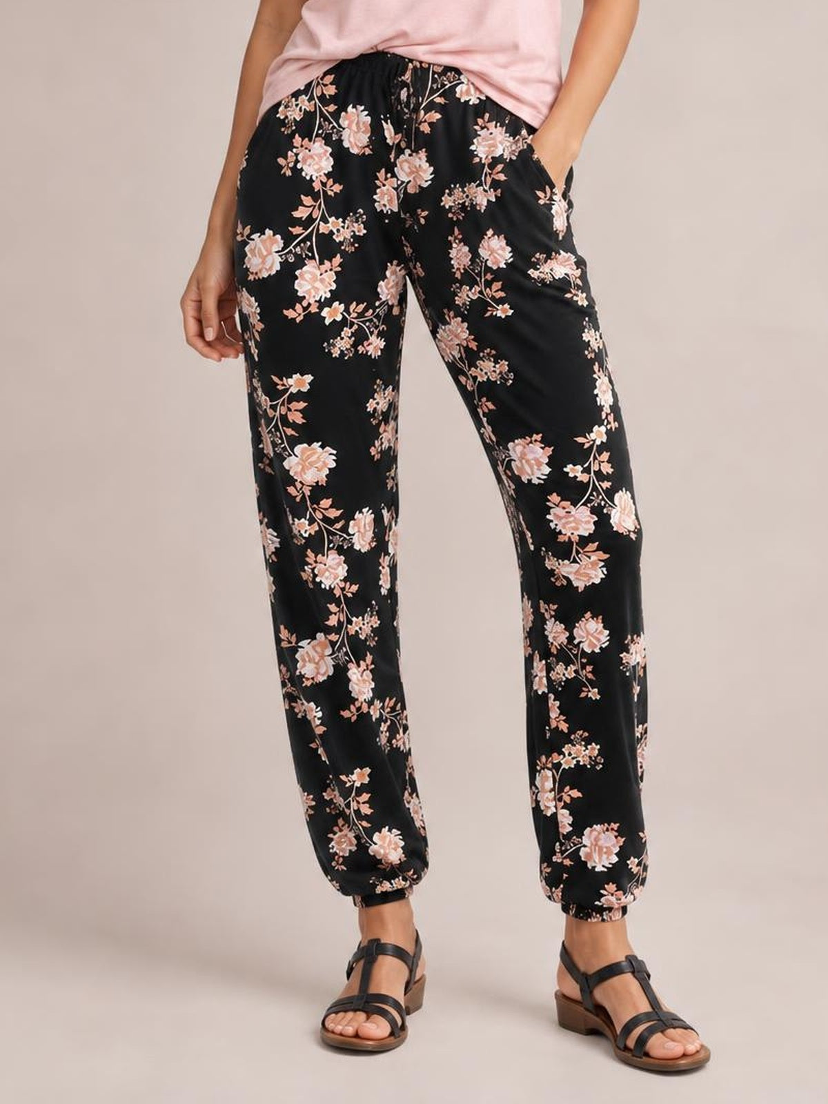 Model wears black drawstring flower pants with pink floral print, side pockets, a relaxed tapered silhouette, elastic cuffs, and hands tucked into the pockets.