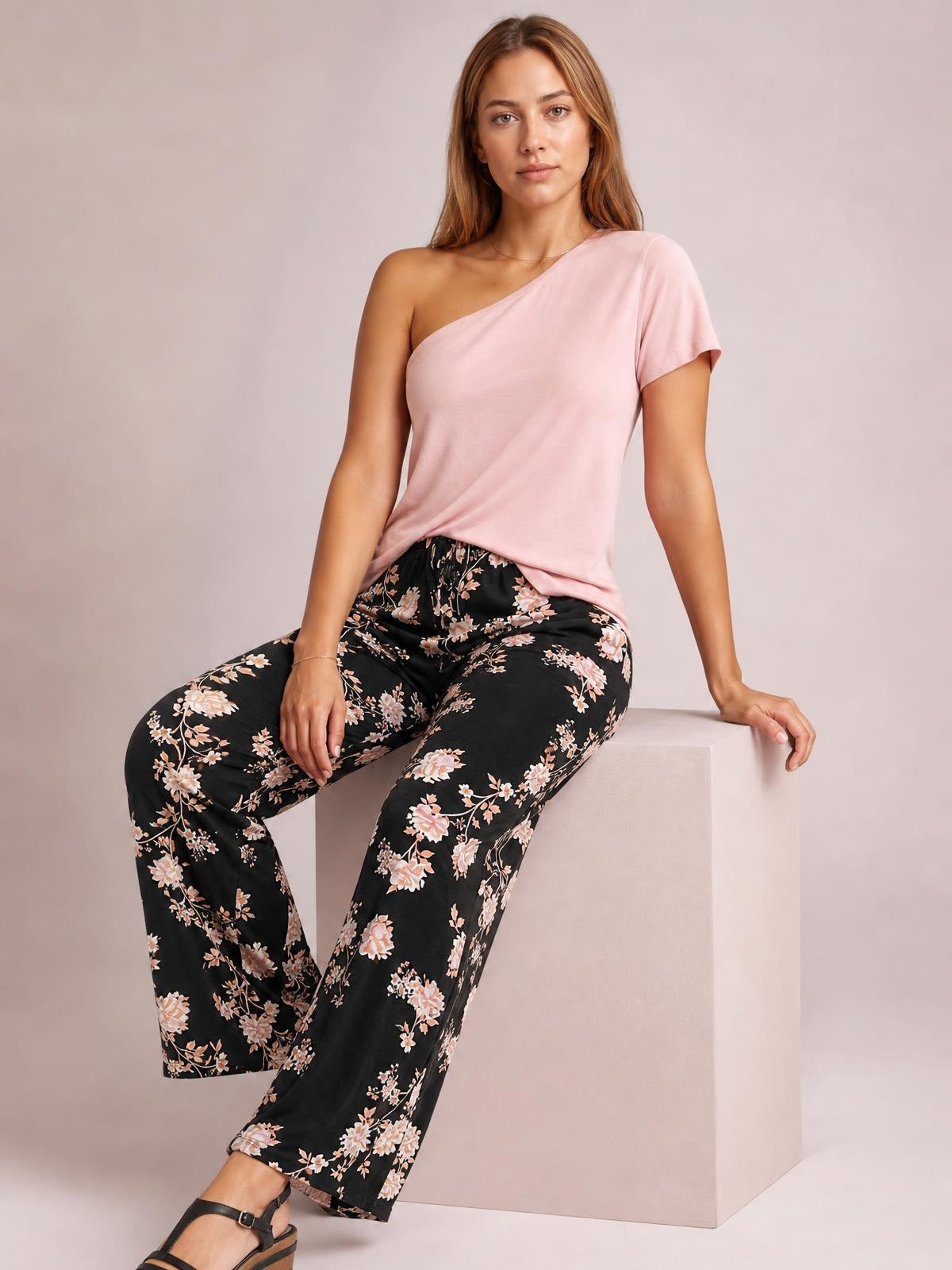 Model seated in black drawstring flower pants with pink floral print, wide-leg silhouette, and visible front pockets.