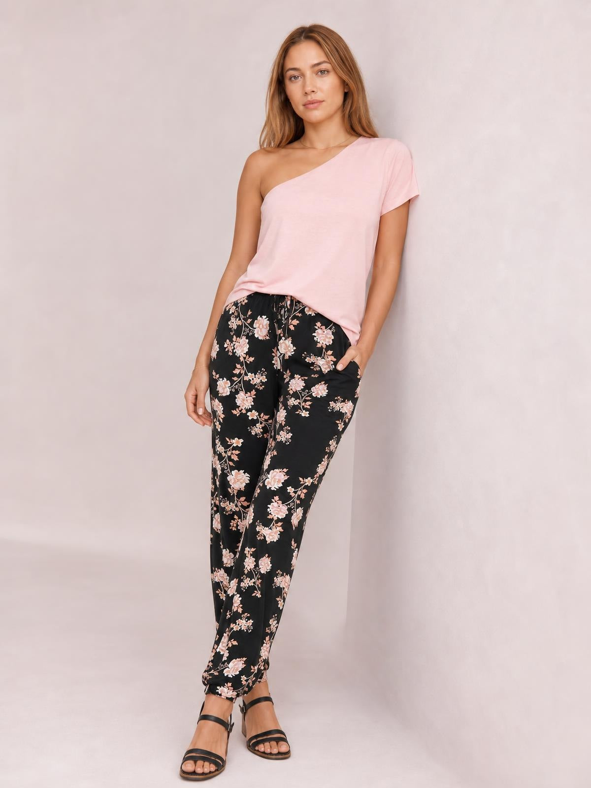 Model poses in black drawstring flower pants with a pink floral print, tapered silhouette, elastic waist, and side pockets.