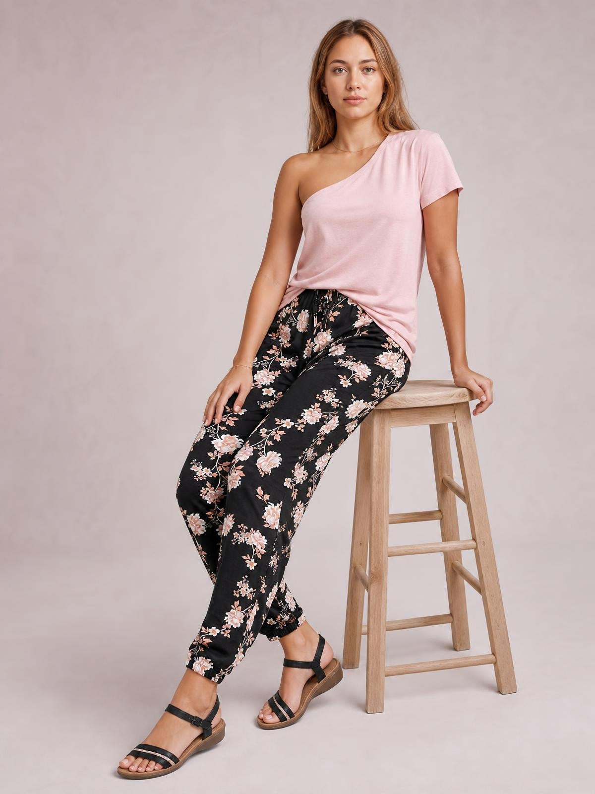 Model seated on a stool wearing black drawstring flower pants with a relaxed tapered silhouette, cropped cuffs, floral print, and visible pockets.