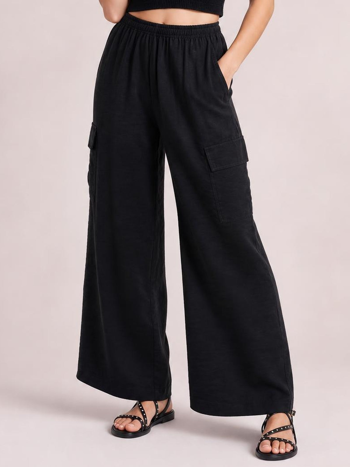 A model stands with one hand in a pocket, wearing black wide-leg utility trousers with a gathered drawstring waist, roomy legs, and cargo pockets.