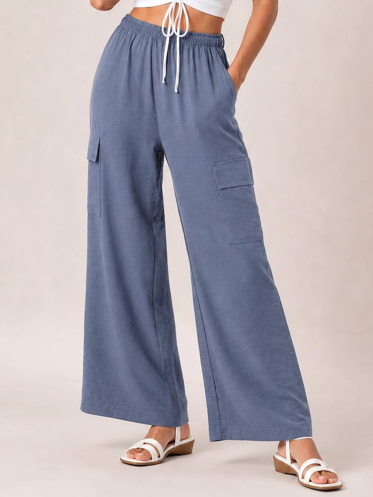 Model wears blue drawstring frilled-waist utility trousers with a wide-leg silhouette, side pockets, and cargo flap pockets, standing with one hand in a pocket.