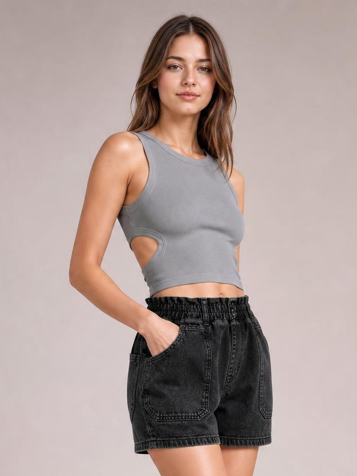 A woman modeling a gray sleeveless crop top with side cutouts and black denim shorts against a plain beige studio background.