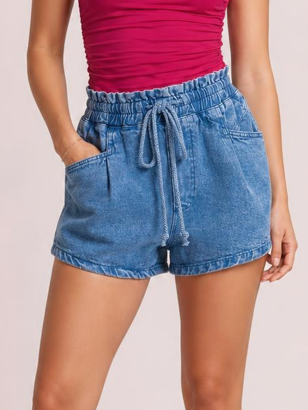 A model wears light blue high-waist denim shorts with a gathered drawstring waistband, front pockets, relaxed silhouette, and cuffed hems, posing with one hand in a pocket.