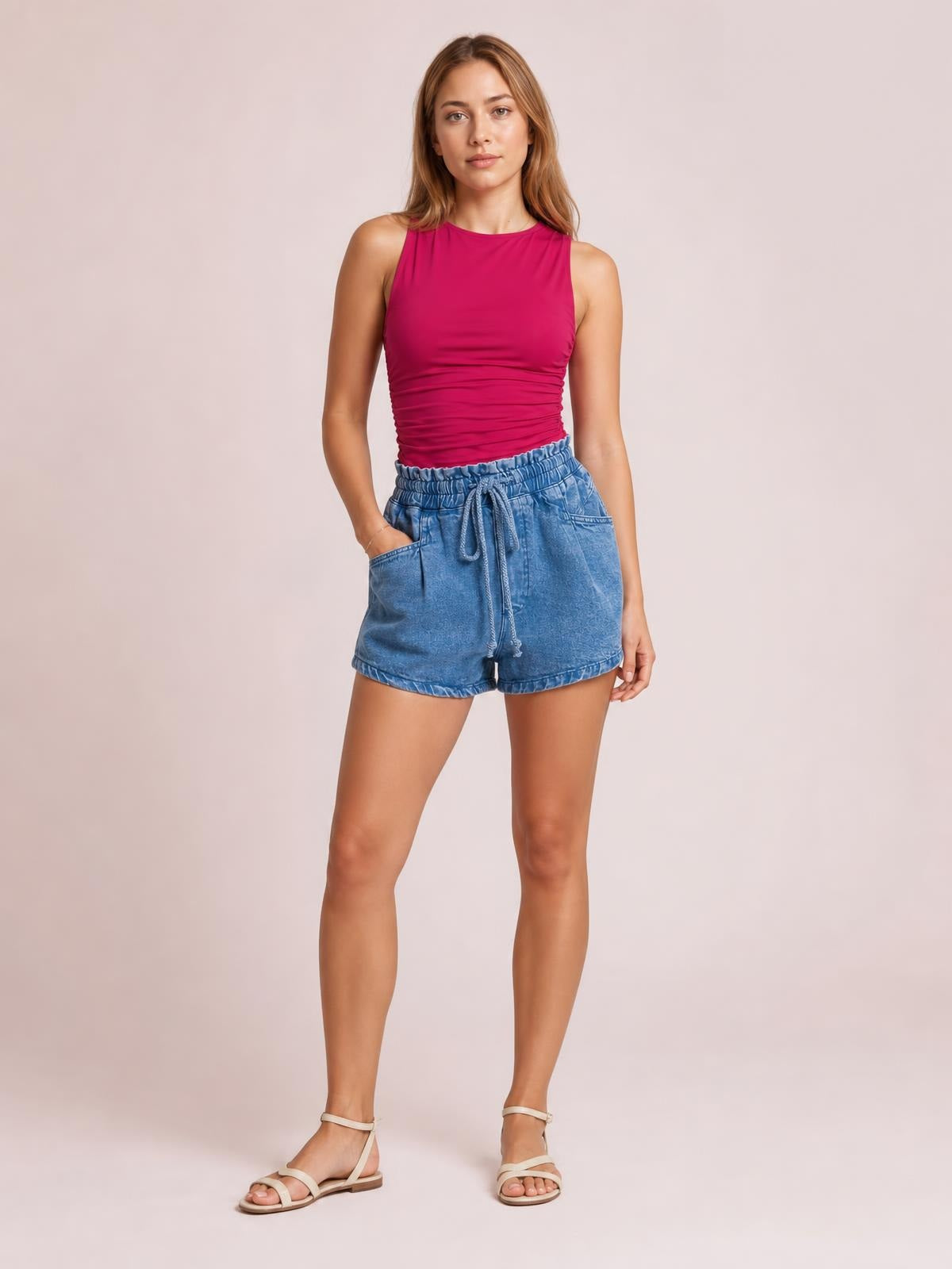 Model standing with one hand in the pocket of blue high-waist denim shorts, featuring a drawstring waist, relaxed silhouette, and cuffed hems.