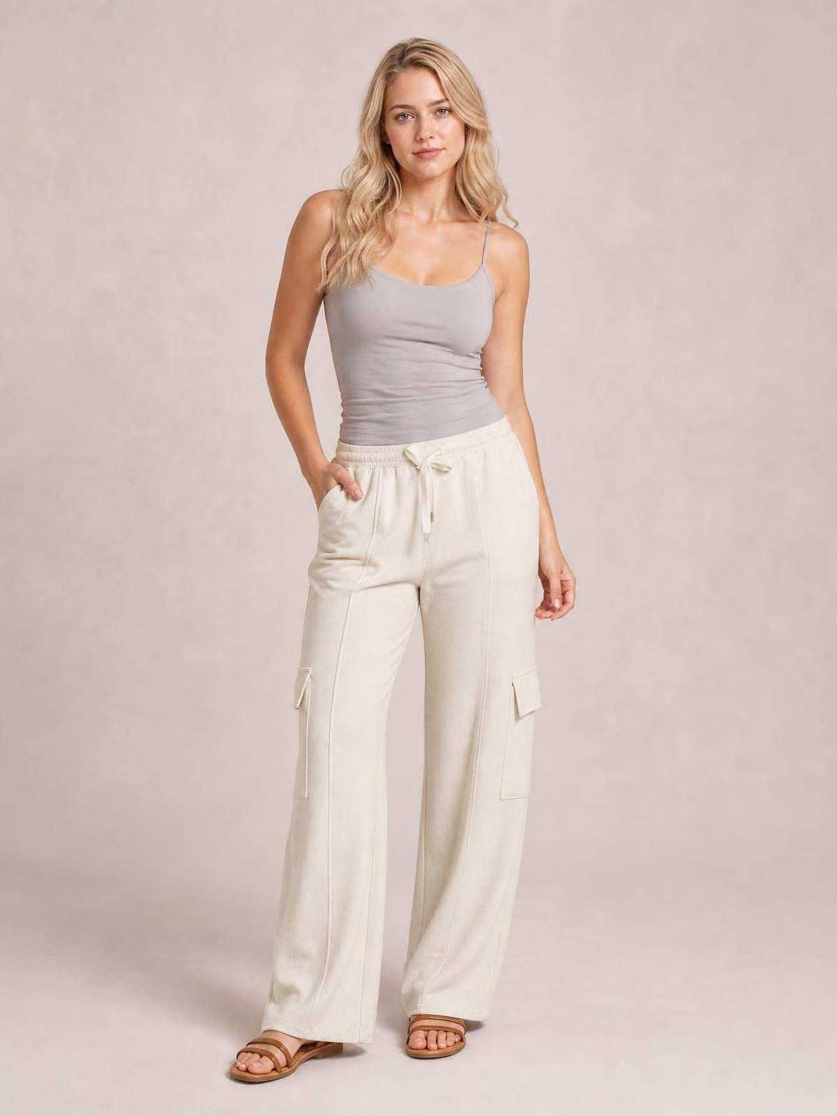 Model poses in cream high-waist drawstring pants with a wide-leg silhouette, side pockets, and flap cargo pockets, placing one hand in a pocket.