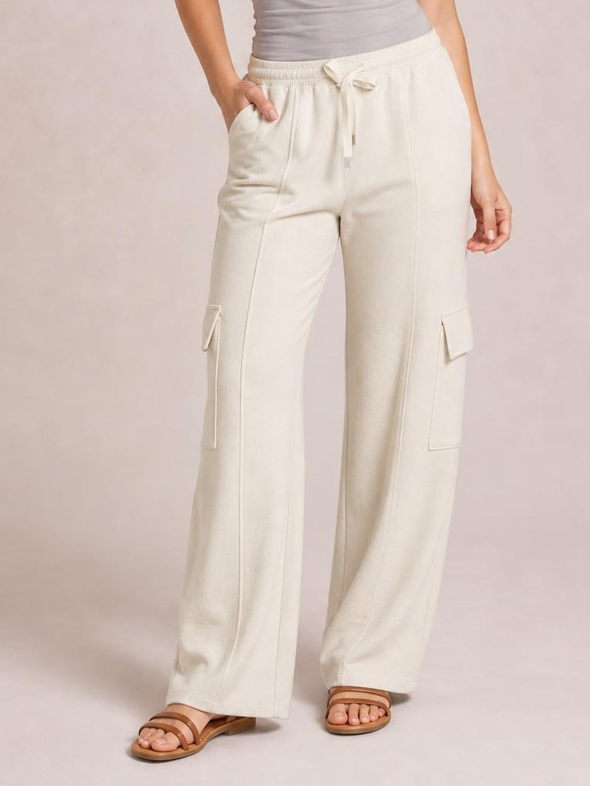 Model poses with one hand in the pocket of cream drawstring high-waist pants featuring wide legs, visible cargo-style pockets, and stitched panel details.