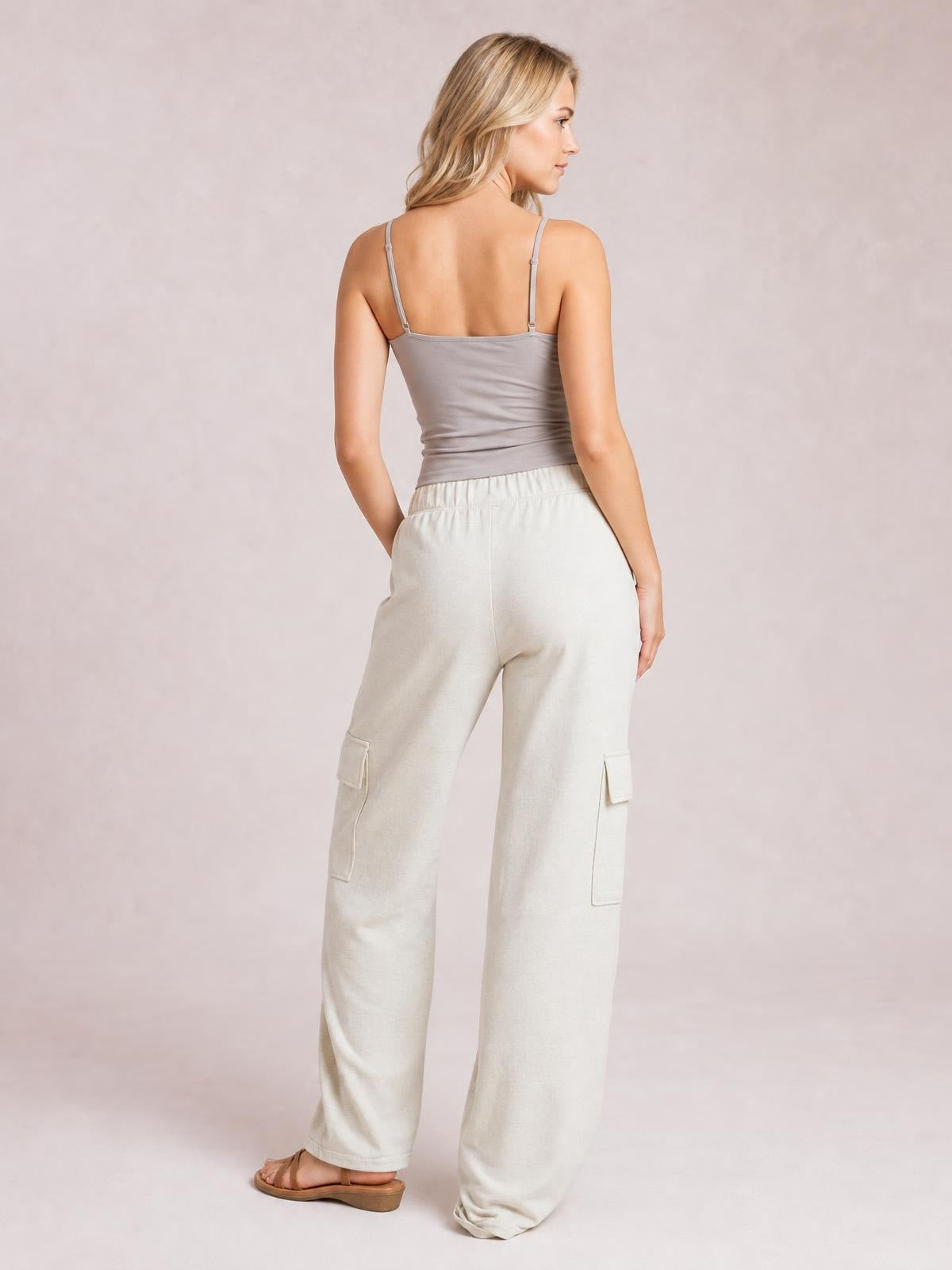 A model poses from the back in light cream high-waist pants with a relaxed wide-leg silhouette, elasticized waistband, and side patch pockets.