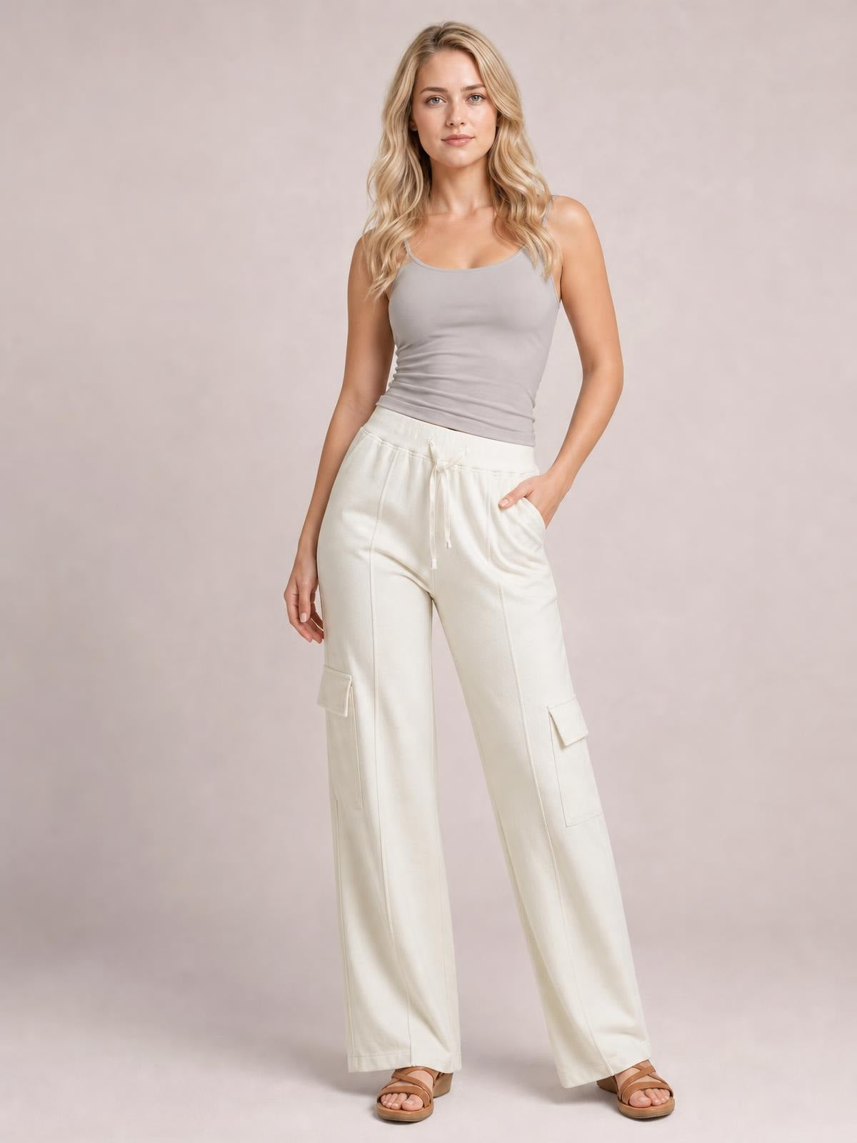 A model stands in cream drawstring high waist pants with wide straight legs, front pockets, and large cargo-style patch pockets, one hand tucked inside.