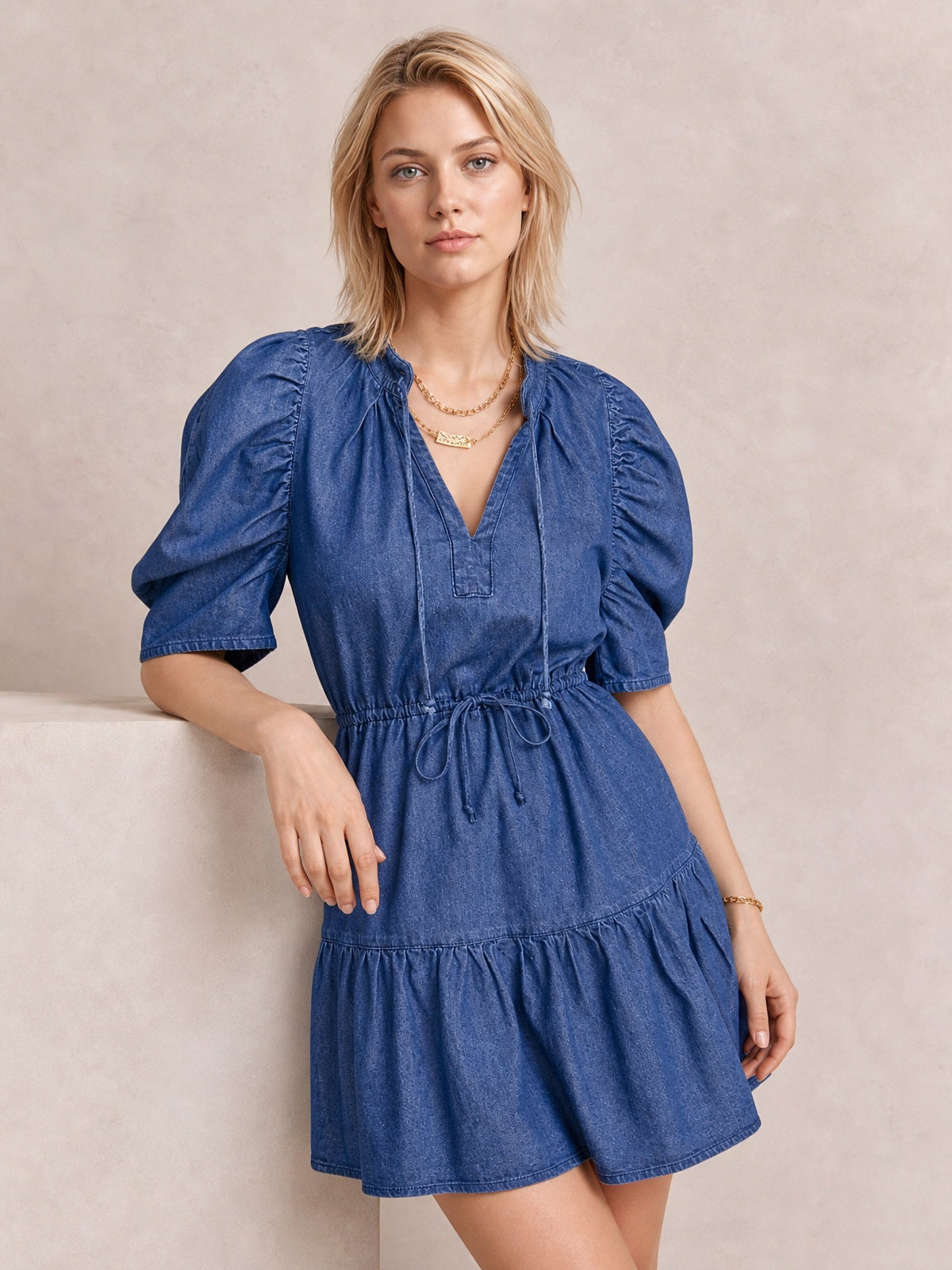 A model wears a navy denim mini dress with puff sleeves, a V-neckline, tied drawstring waist, and gathered seam creating a softly flared skirt silhouette.