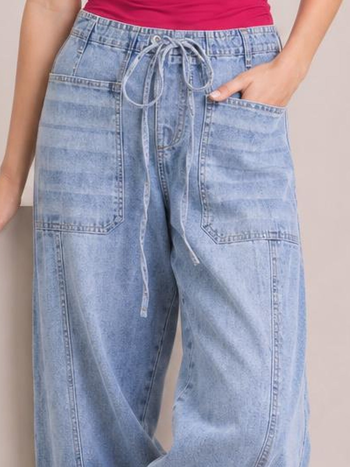 Model wearing light blue drawstring jeans with large patch pockets, a relaxed wide-leg silhouette, and a tied waist cord, with one hand in a pocket.