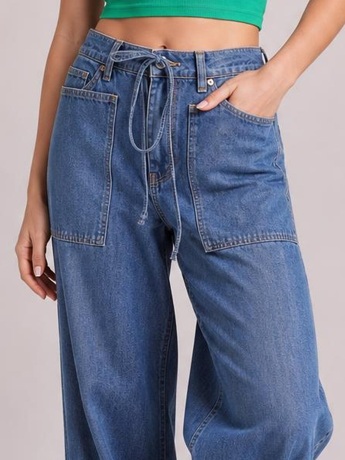 A model showcases blue drawstring jeans with a relaxed wide-leg silhouette, oversized patch pockets, belt loops, and one hand tucked into a pocket.