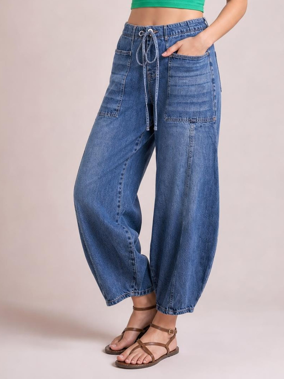 Model wears blue drawstring jeans with large patch pockets, a relaxed wide-leg silhouette, cropped ankle-length hems, and one hand tucked into a pocket.