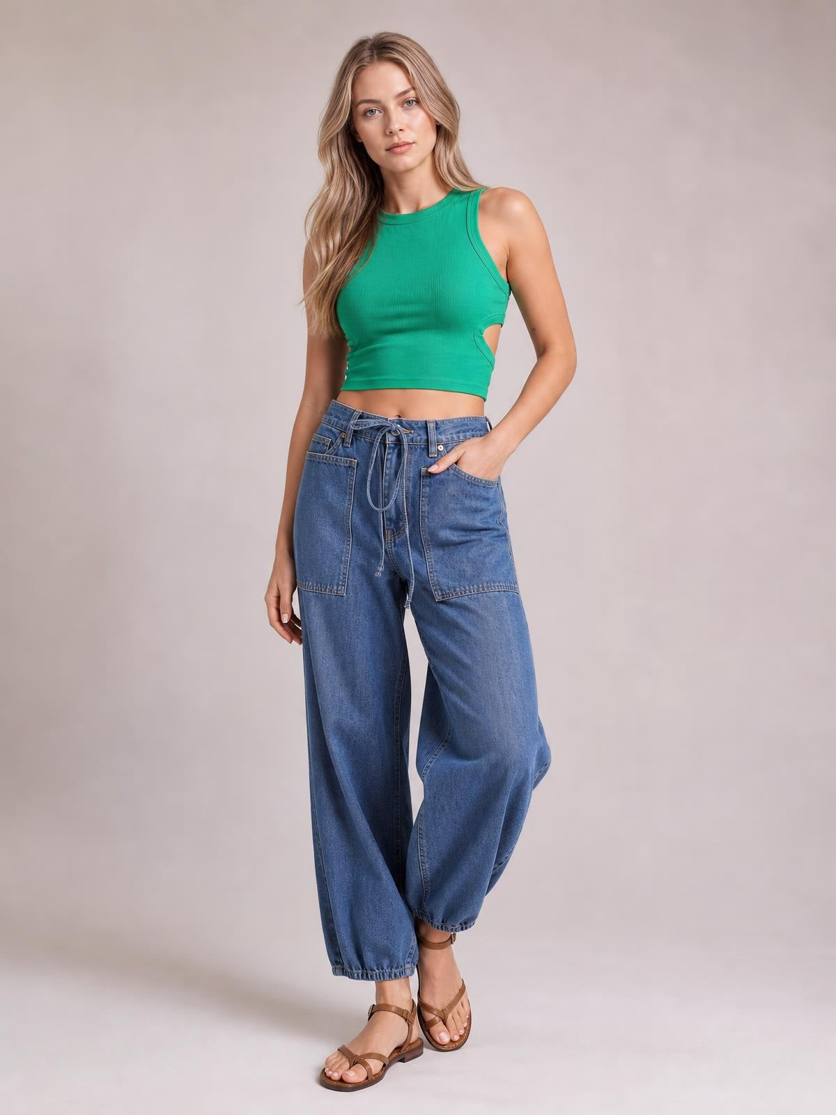 Model poses with one hand in the pocket, wearing blue drawstring jeans with front patch pockets, a relaxed wide-leg silhouette, and cropped hems.