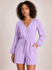 Model wearing a lavender drawstring long sleeve romper with a wrap-style neckline, elastic cuffs, relaxed silhouette, and tie waist, posing with one hand in a pocket.