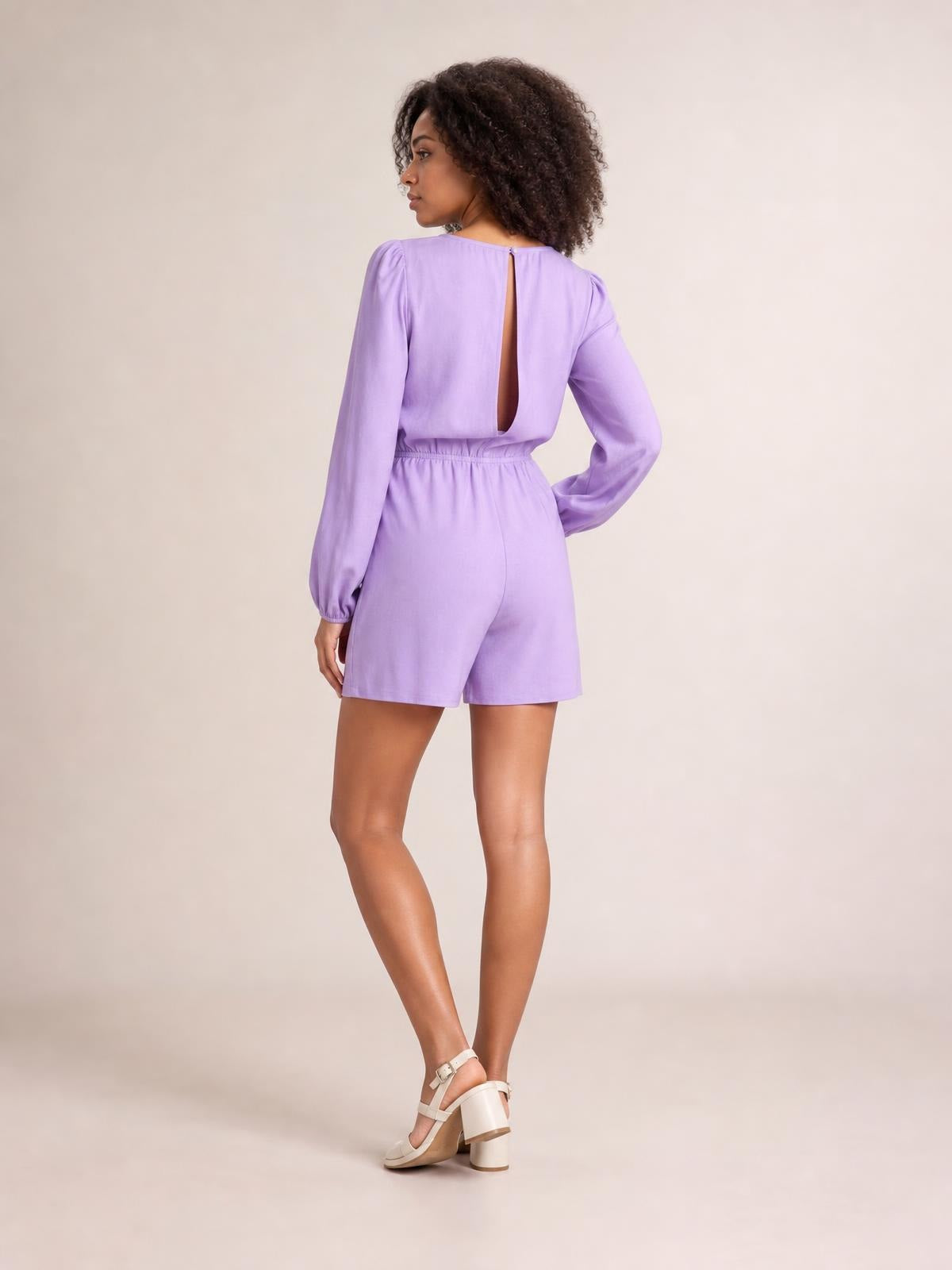 Model poses in a lavender long sleeve romper with a drawstring waist, relaxed silhouette, gathered cuffs, and tailored shorts.