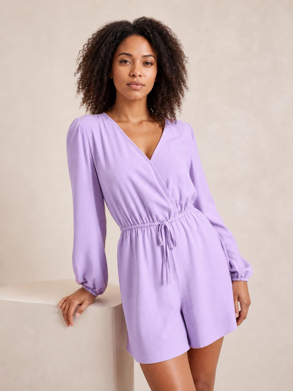 Model leans against a ledge wearing a lavender drawstring long sleeve romper with a wrap-style neckline, relaxed fit, and breezy shorts silhouette.