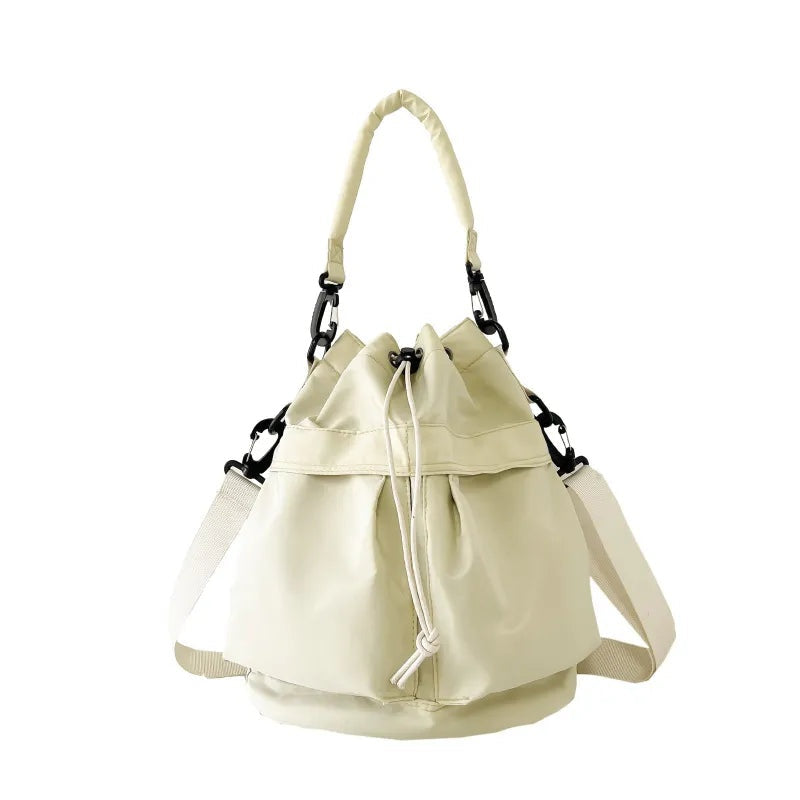 Cream nylon bucket bag with a softly gathered drawstring opening, structured base, top handle, and adjustable side straps for versatile carrying.