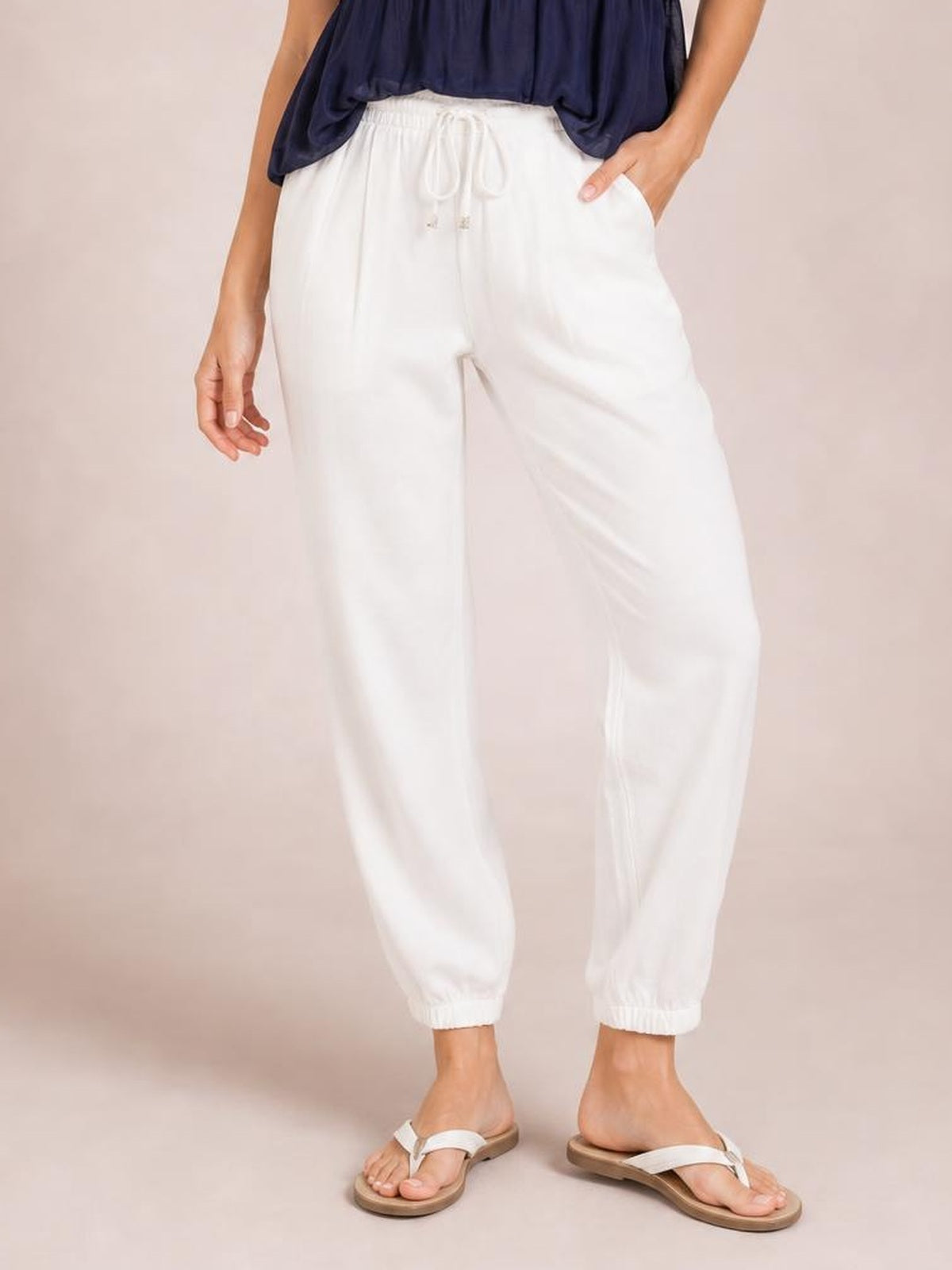A model stands in white cropped joggers with a gathered paperbag waist, adjustable drawstring, relaxed tapered legs, and elasticized ankle cuffs.