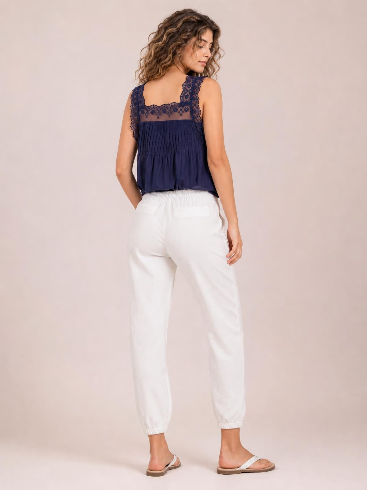 A model wears white cropped joggers with a gathered paperbag waist, relaxed tapered legs, and elastic ankle cuffs in a back-facing pose.
