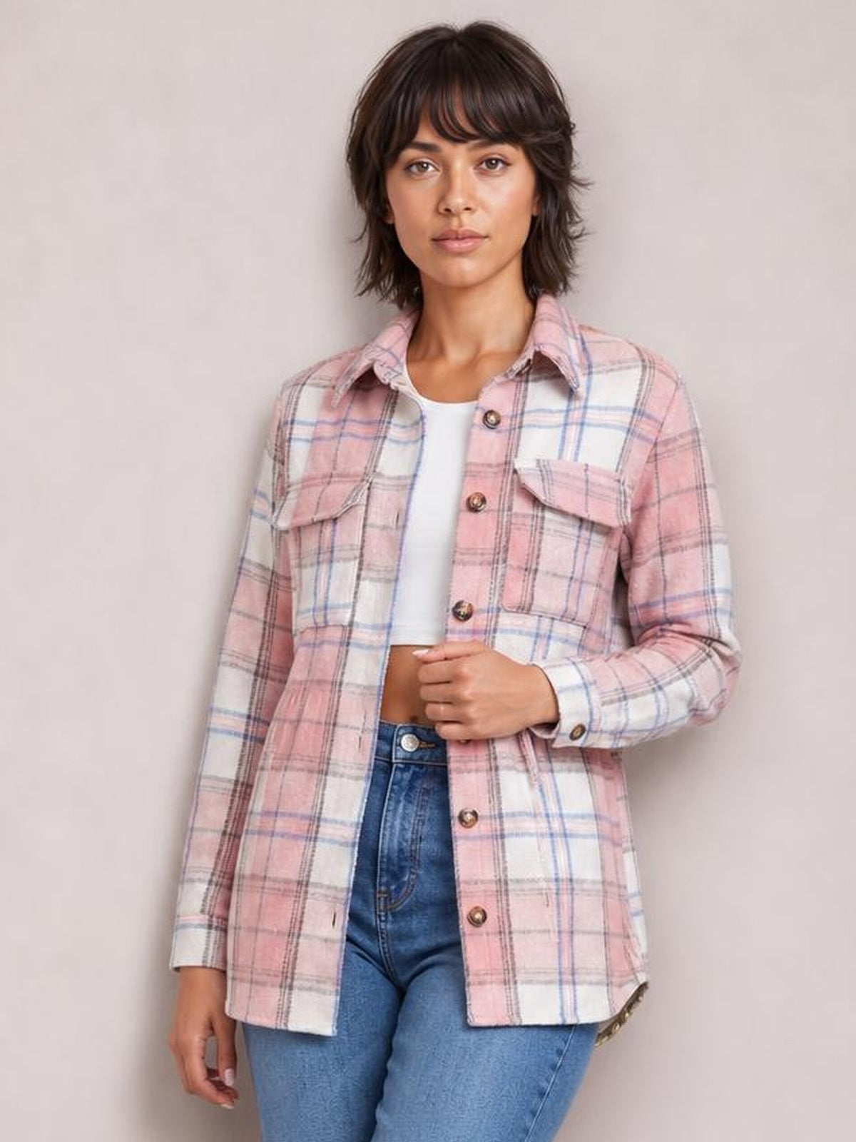 Model poses in a pink plaid button-up jacket with a collared silhouette, chest flap pockets, long sleeves, and front buttons over a white top.